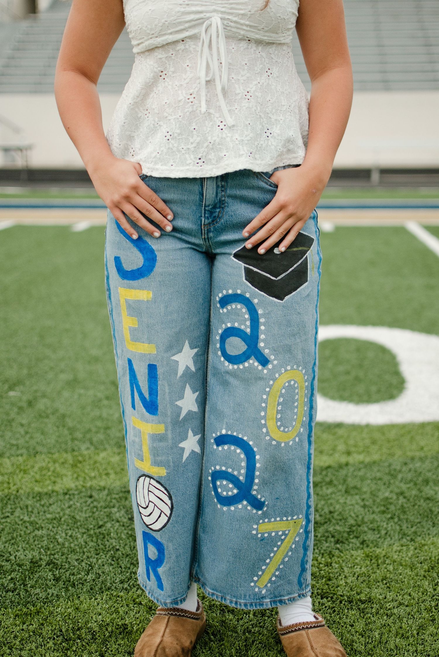Close-up of wide-leg jeans painted with SENIOR 2027, graduation cap, volleyball, and stars design.