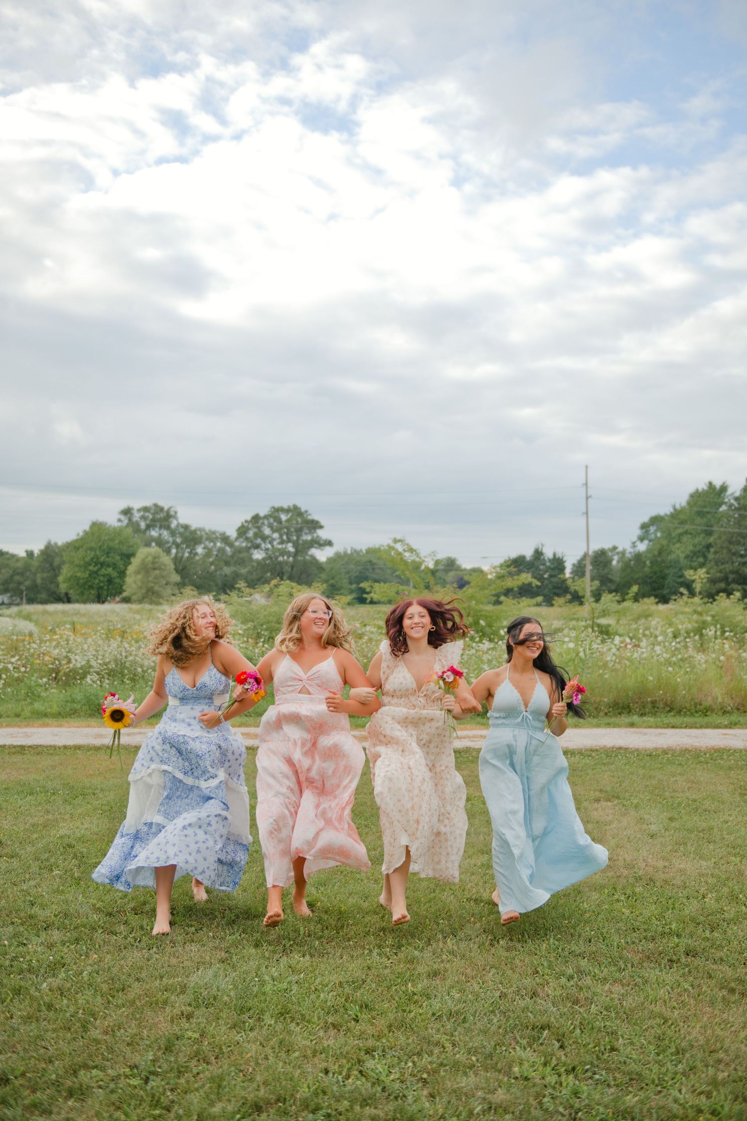 Four bridesmaids in pastel maxi dresses running and laughing across a green field with bouquets.