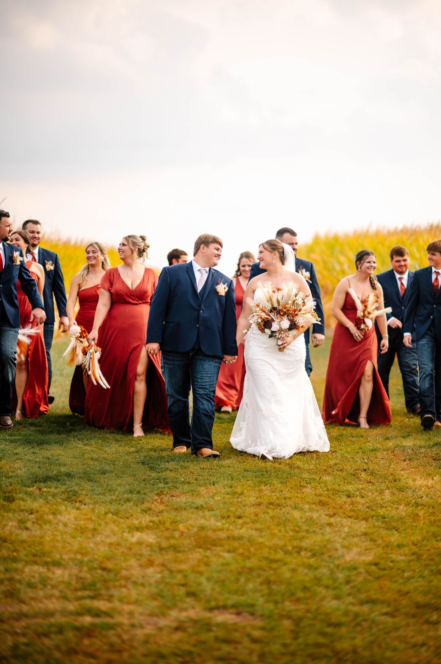 Wedding party walks through a grassy field with bridesmaids in coral dresses.