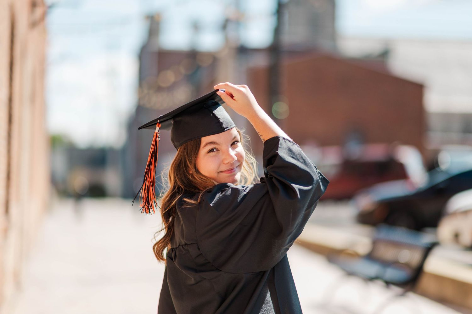 Graduate in black cap and gown smiling over shoulder while adjusting mortarboard in urban setting.