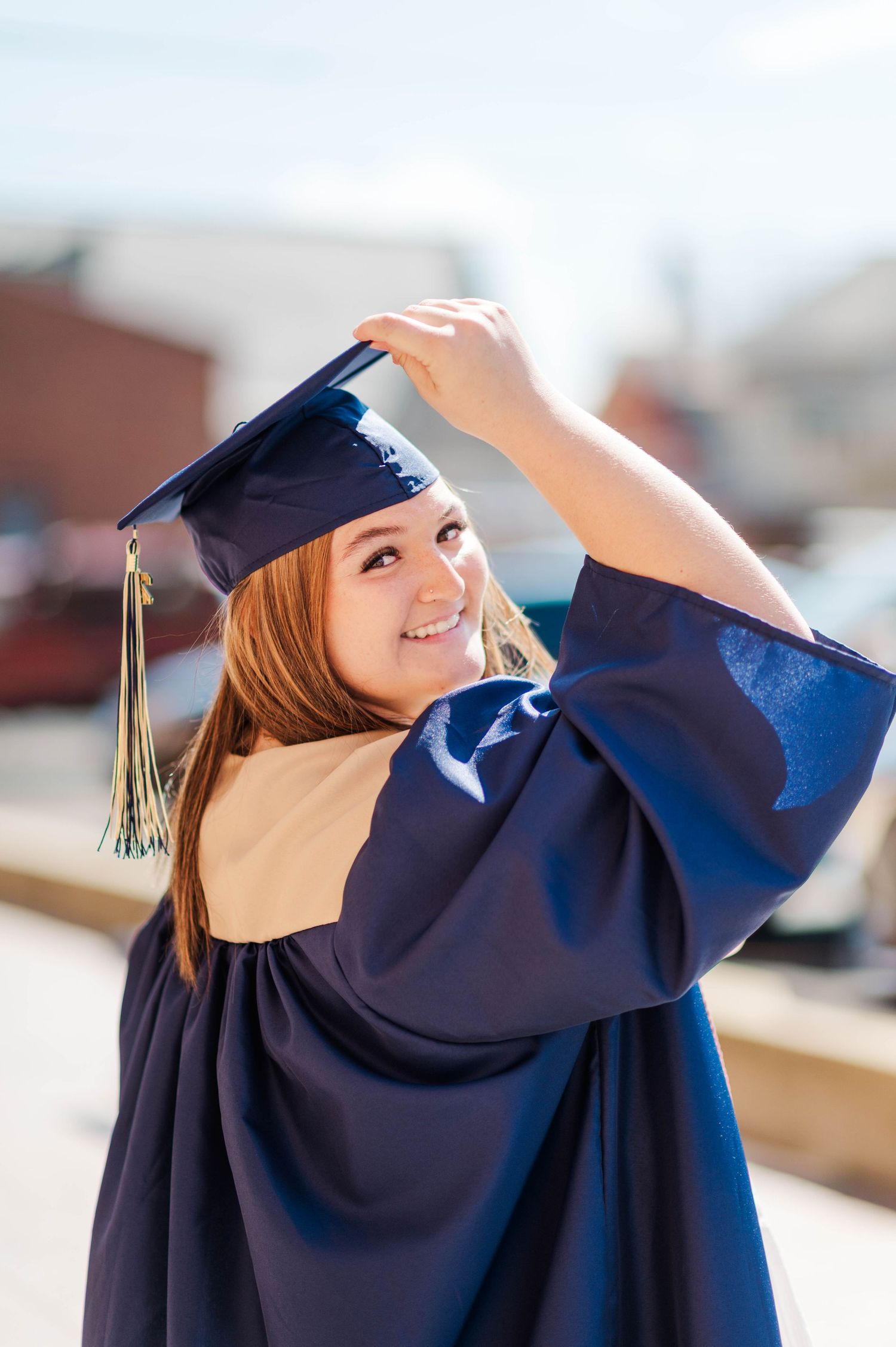 Redhead graduate in navy cap and gown smiling over shoulder while adjusting mortarboard outdoors.