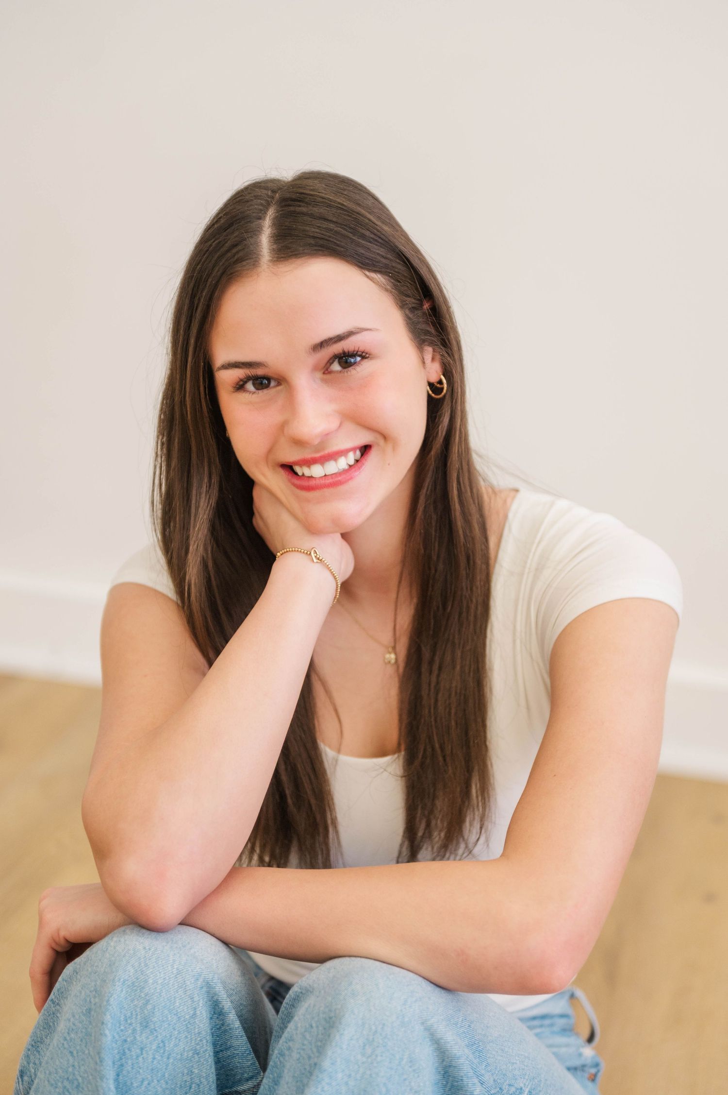 Smiling young woman with long brown hair sitting on floor in white tee and jeans, resting chin on hand.