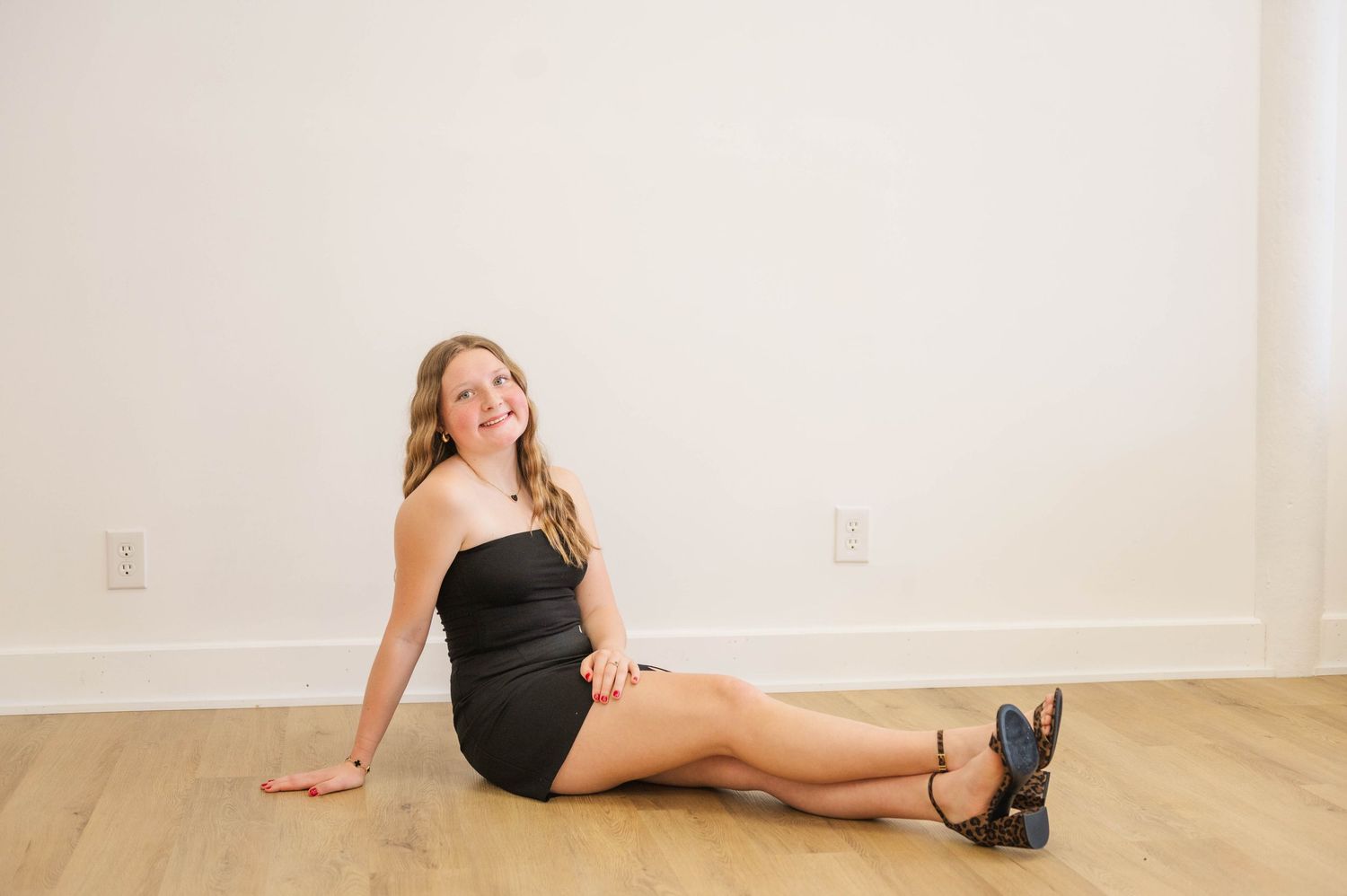Teen girl in black strapless mini dress and heels sitting on hardwood floor, smiling.