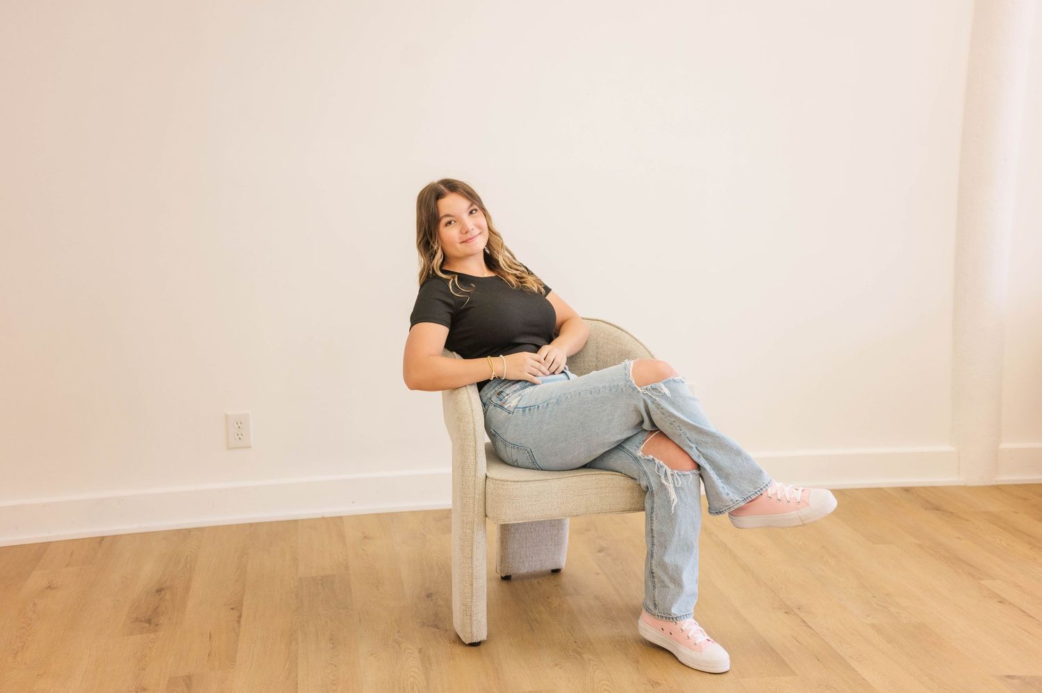 Young woman in black top and ripped jeans sitting relaxed on a chair, wearing pink sneakers, on hardwood floor.