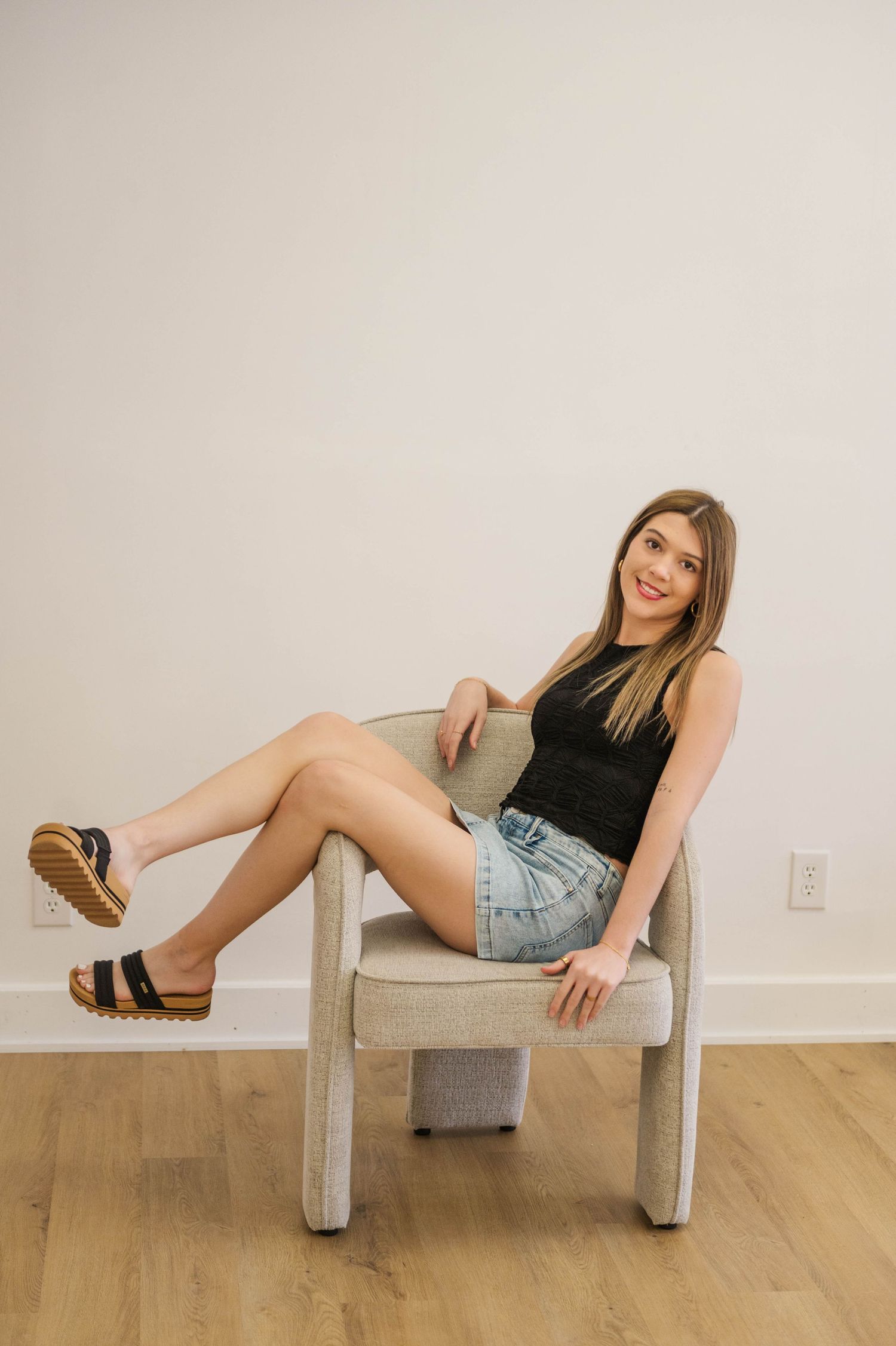 Smiling woman in black sleeveless top and denim mini skirt relaxes in chair with legs crossed.