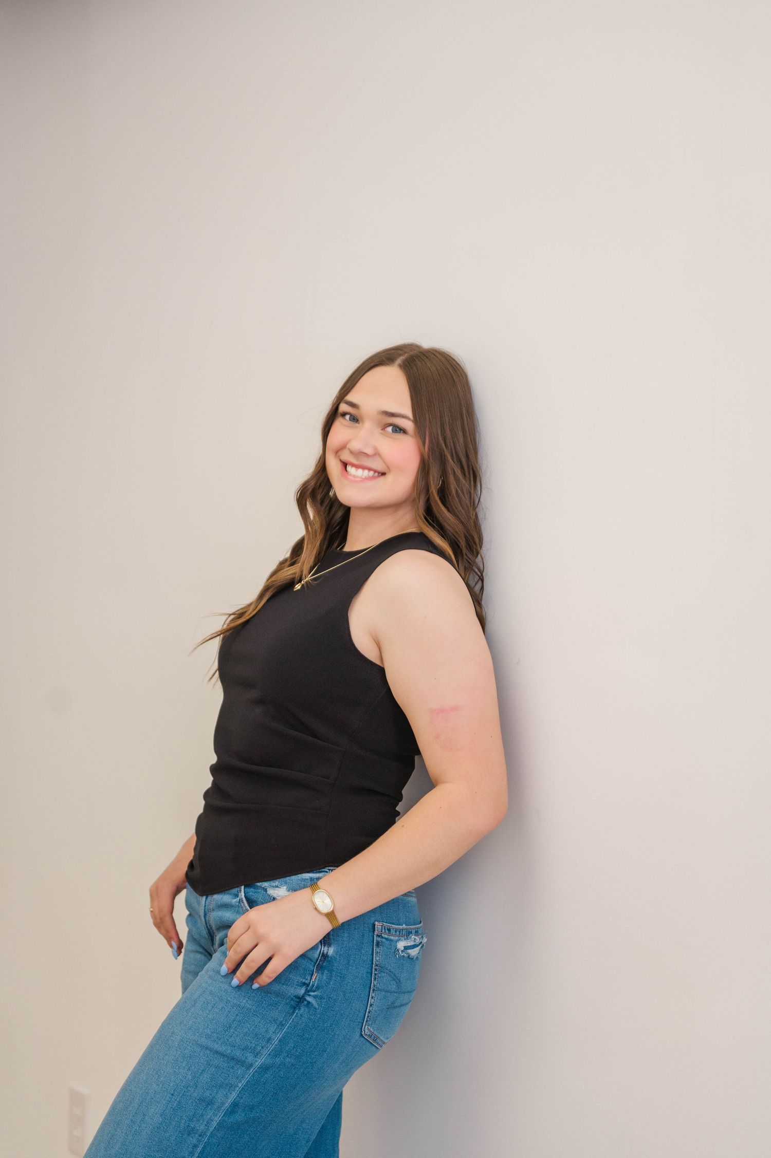 Smiling young woman in black tank top and blue jeans leans against white wall in casual pose.