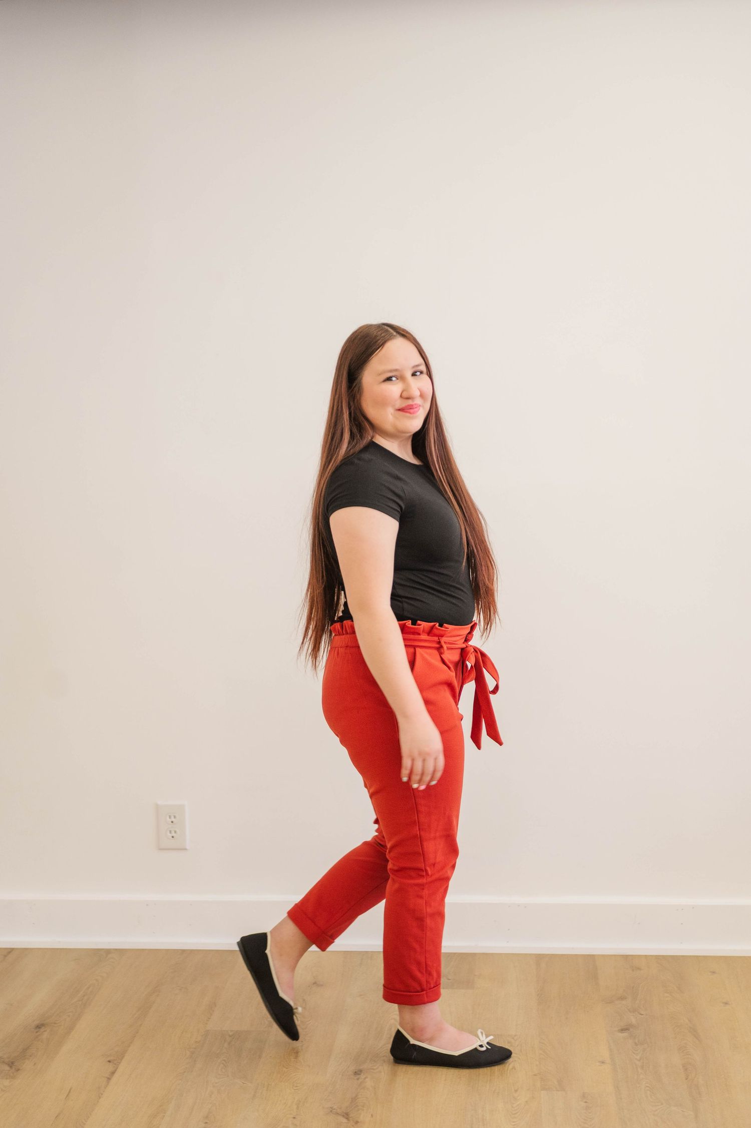 Brunette woman in black top and red pants walking in profile view in a white studio.