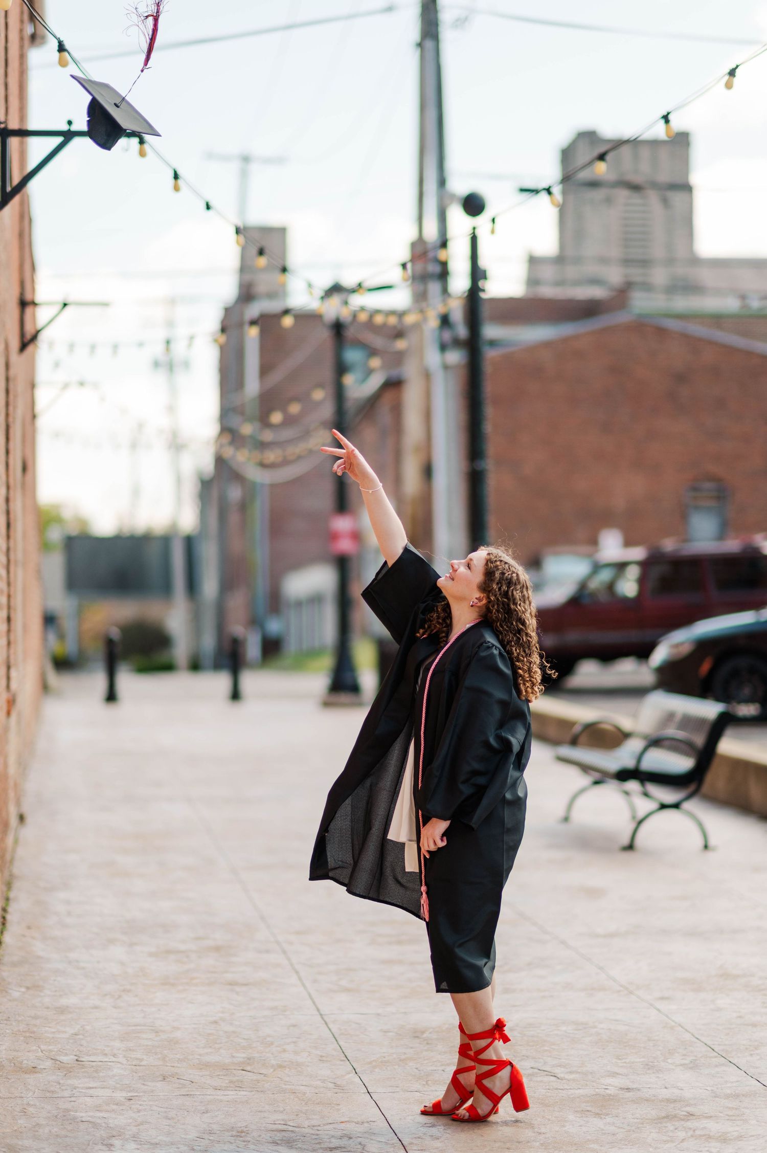 Graduate in black gown tossing cap in air on urban sidewalk with string lights.