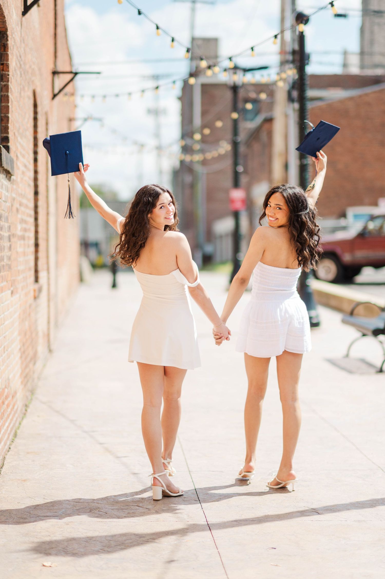 Two graduates in white dresses holding navy mortarboards in the air while walking hand in hand.