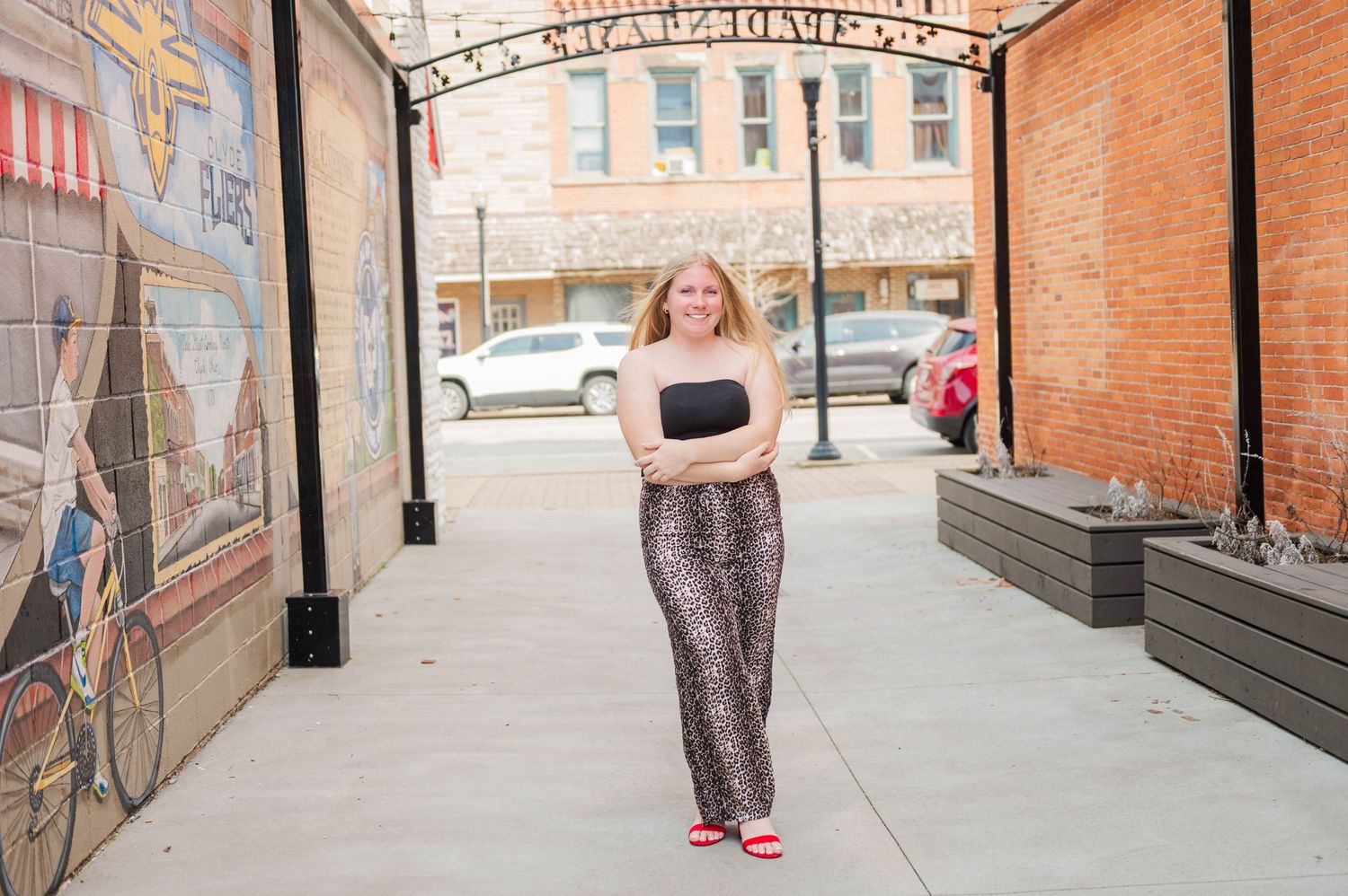 Young woman in black tube top and leopard print pants poses with arms crossed on outdoor patio.