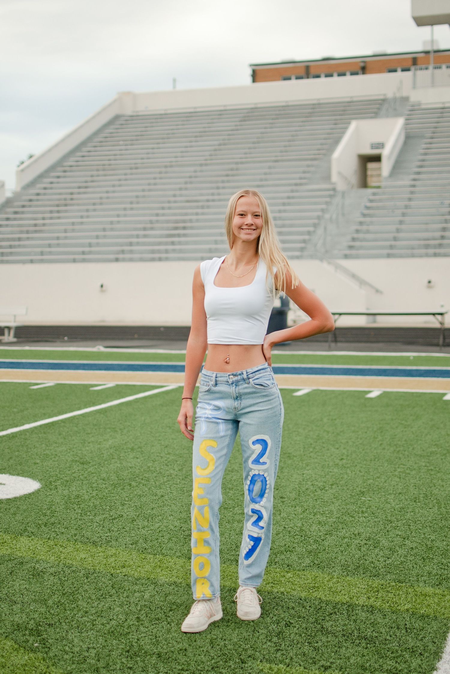 Smiling blonde senior girl stands on football field wearing white crop top and custom painted Class of 2027 jeans.