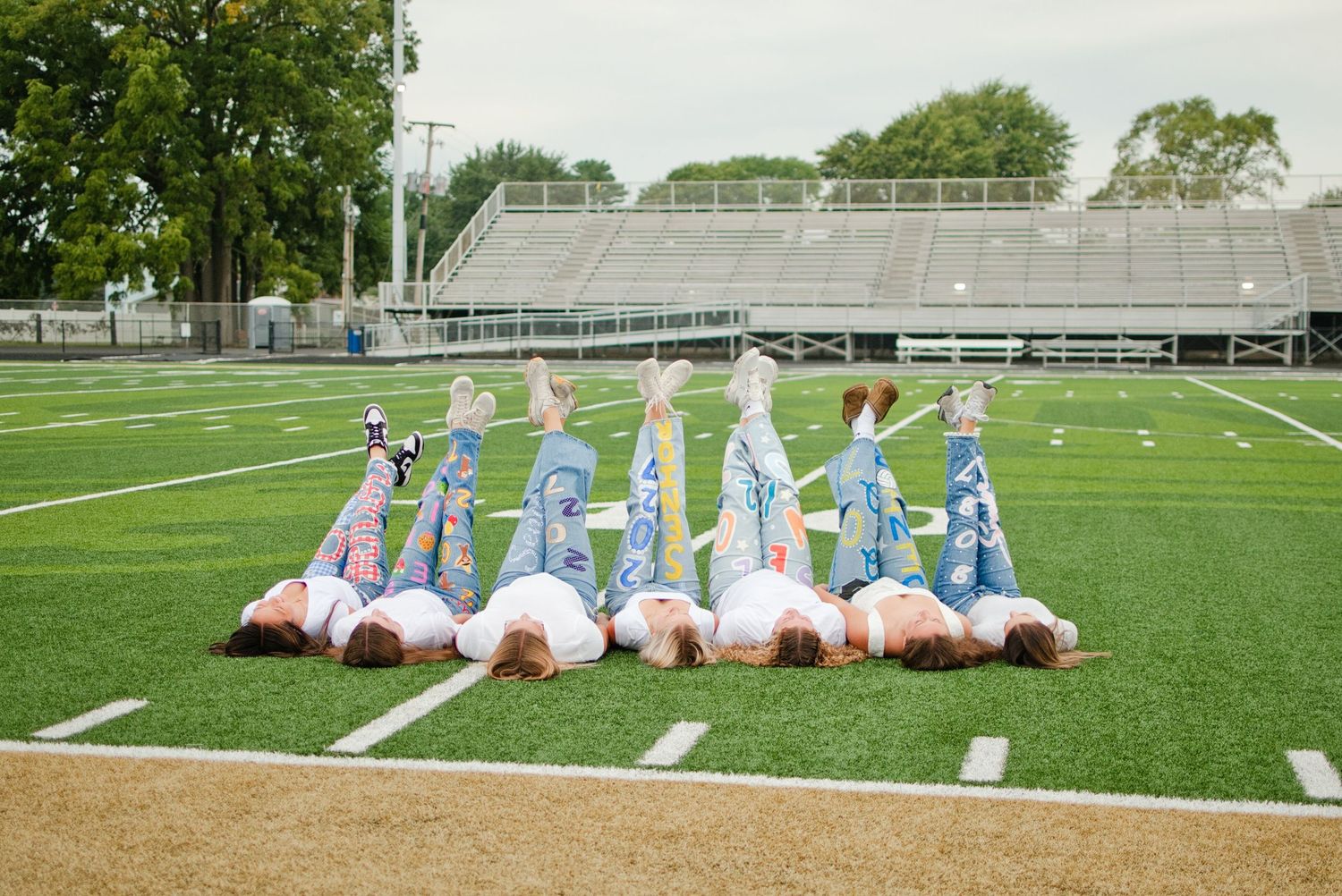 Group of girls lying on football field with legs raised in the air, wearing jeans, with stadium bleachers behind.