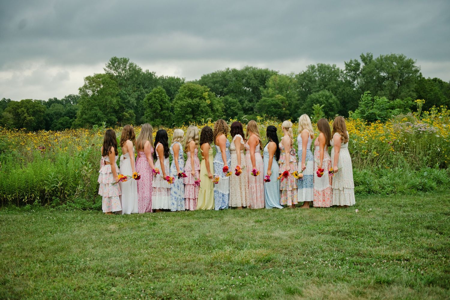A group of bridesmaids in colorful floral dresses stand together in a lush green field holding bouquets.