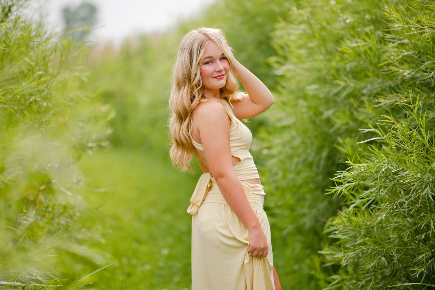 Blonde woman in yellow maxi dress posing among lush green foliage outdoors.