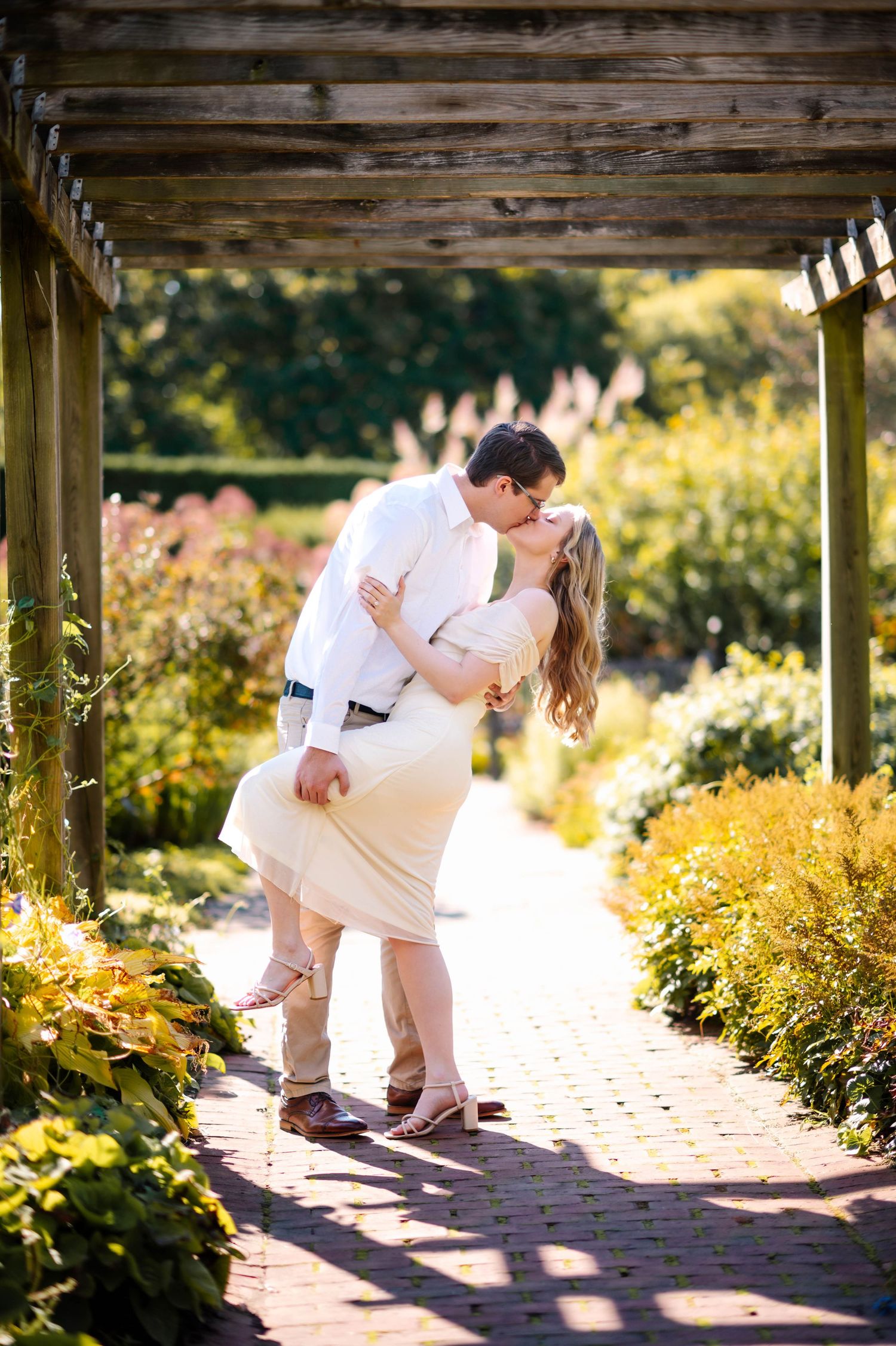A romantic couple sharing a passionate kiss under a wooden pergola surrounded by blooming gardens.