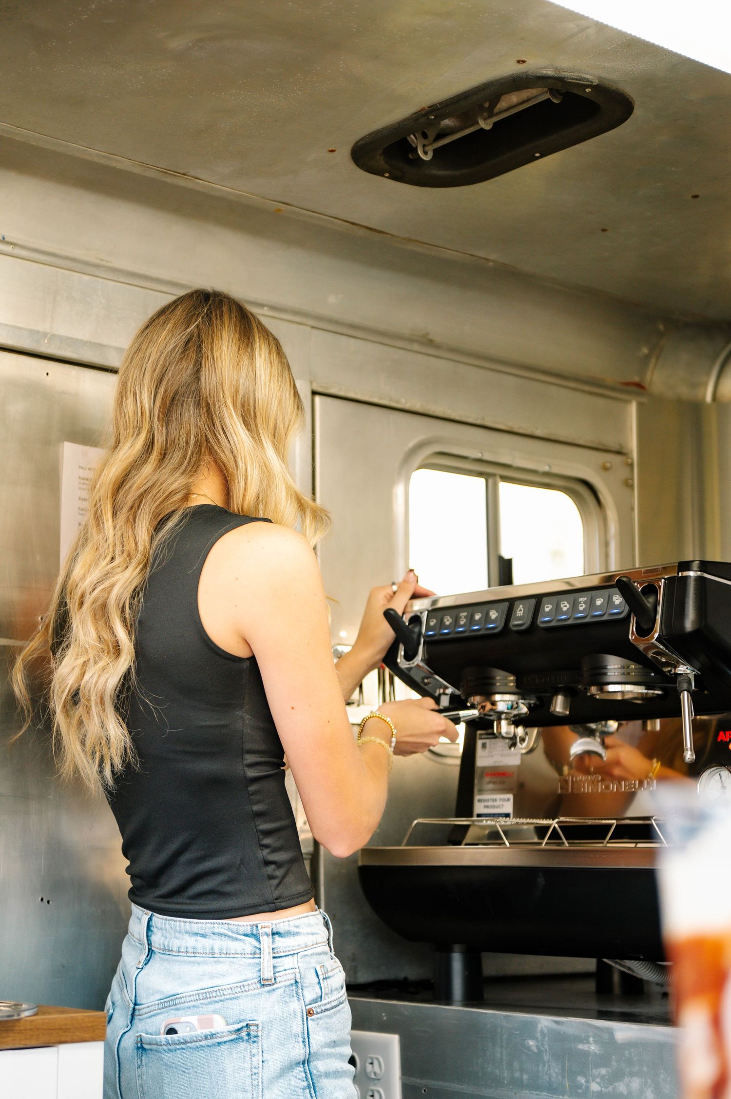 Person in black tank top and jeans working behind a coffee counter.