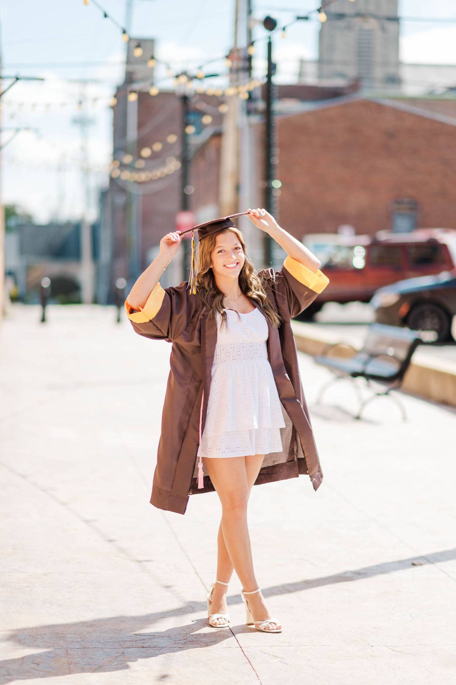 Smiling graduate in brown and gold cap and gown holding mortarboard on sunny urban sidewalk.