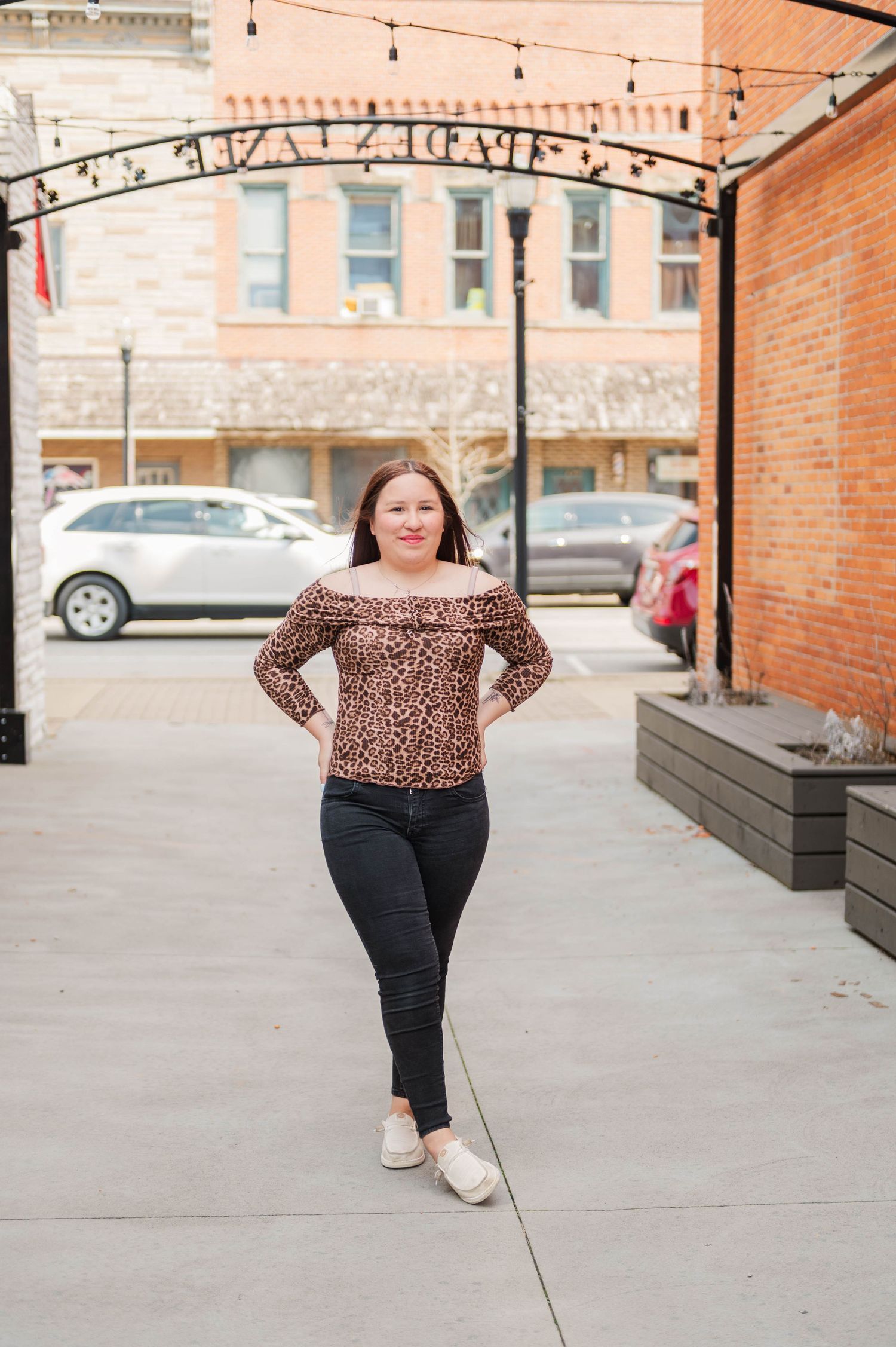 Woman in off-shoulder leopard print top and black jeans poses under decorative arch in downtown area.
