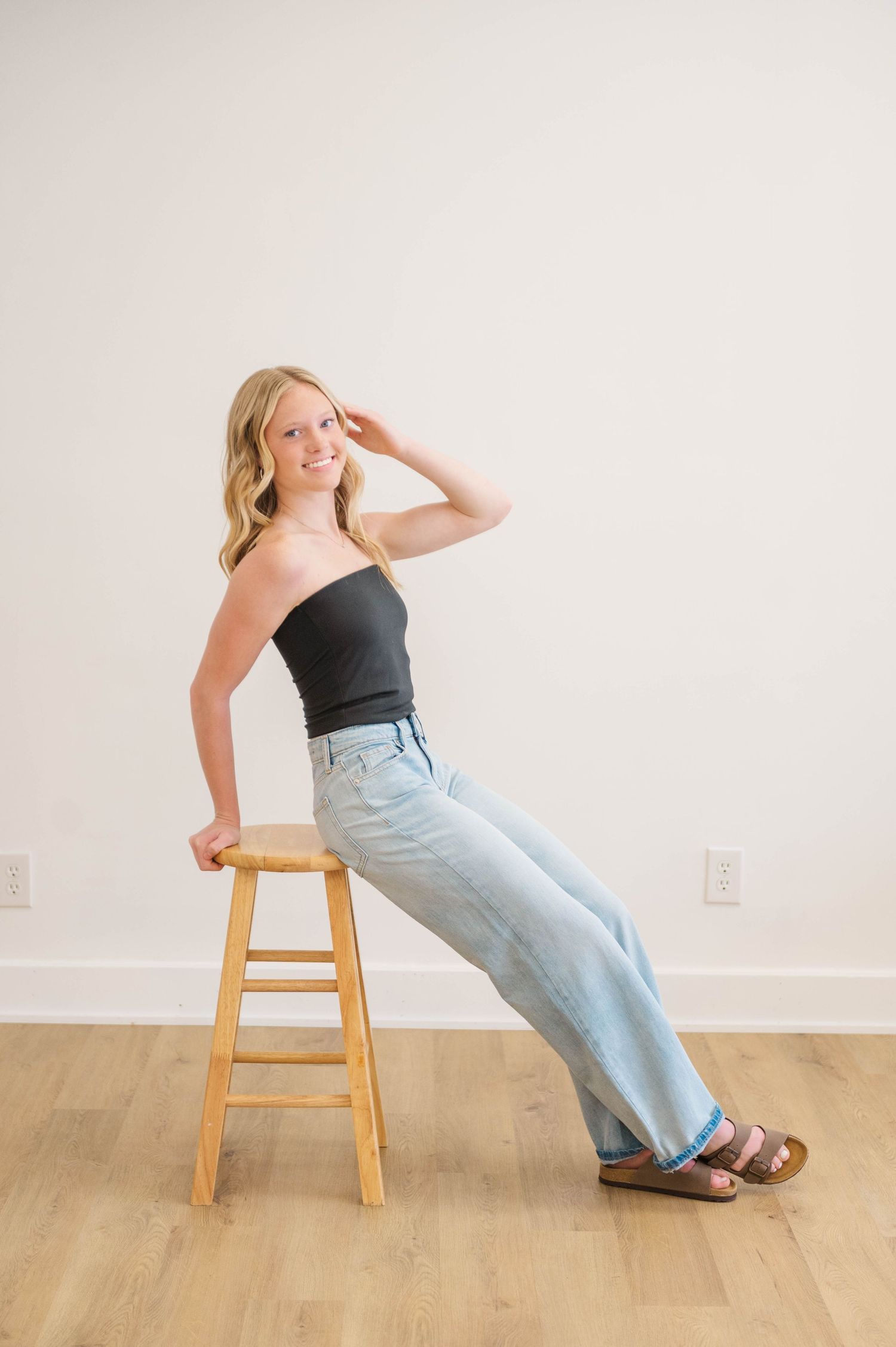 Blonde woman in black tube top and wide-leg jeans leaning on wooden stool, smiling in bright minimalist room.