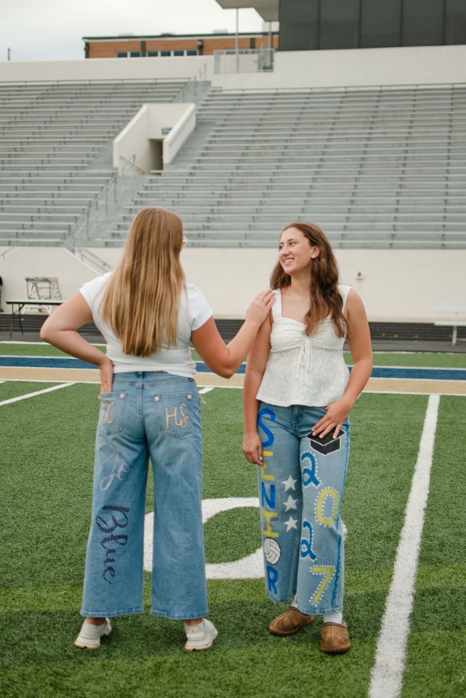 Two teenage girls in decorated senior jeans stand on a football field, one showing off painted denim with school letters.