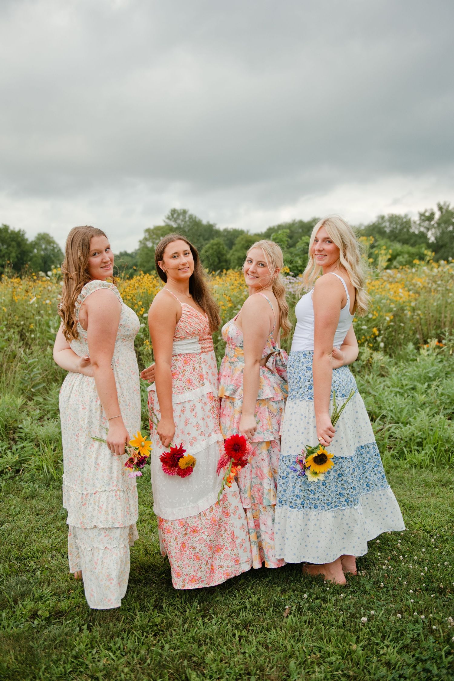 Four women in floral maxi dresses holding colorful bouquets pose in a sunflower field.
