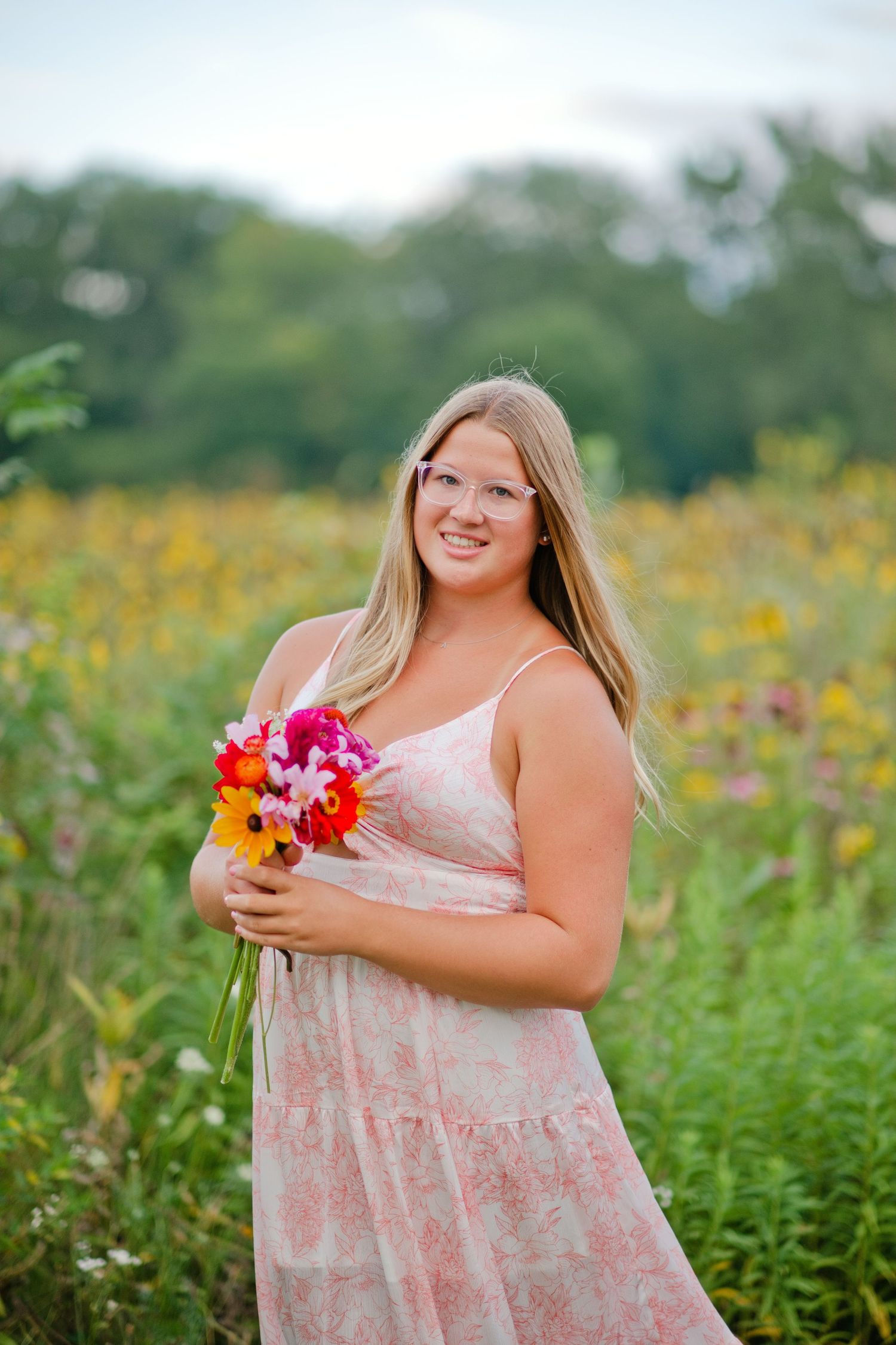 Smiling blonde woman in pink floral sundress holding colorful bouquet in vibrant wildflower field.