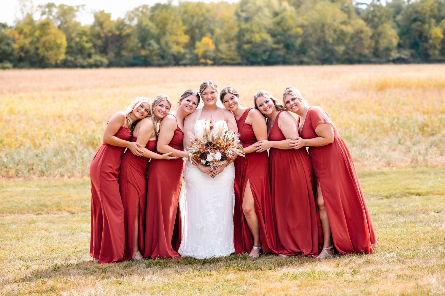 Wedding party in red dresses poses creatively on grassy field during autumn.