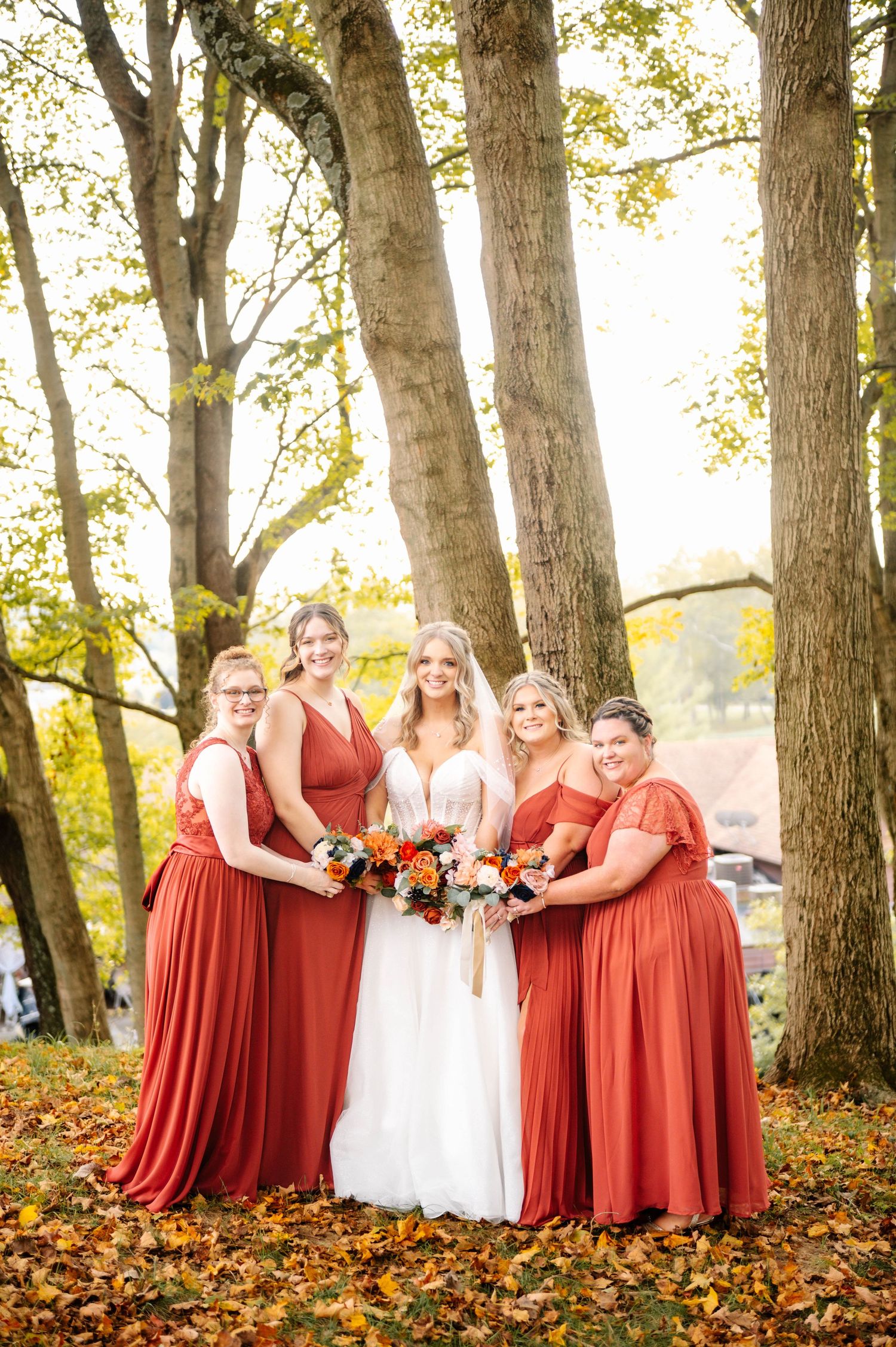 Bridal party in rust-colored dresses poses among fall foliage under tall trees.