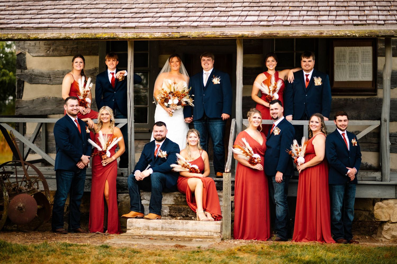 Wedding party in red dresses and navy suits pose together in front of rustic building.