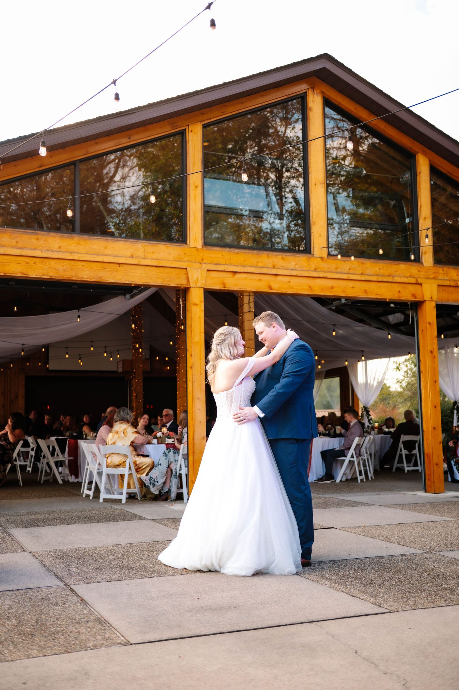 Wedding couple shares a romantic dance under wooden beams and string lights at an outdoor venue.