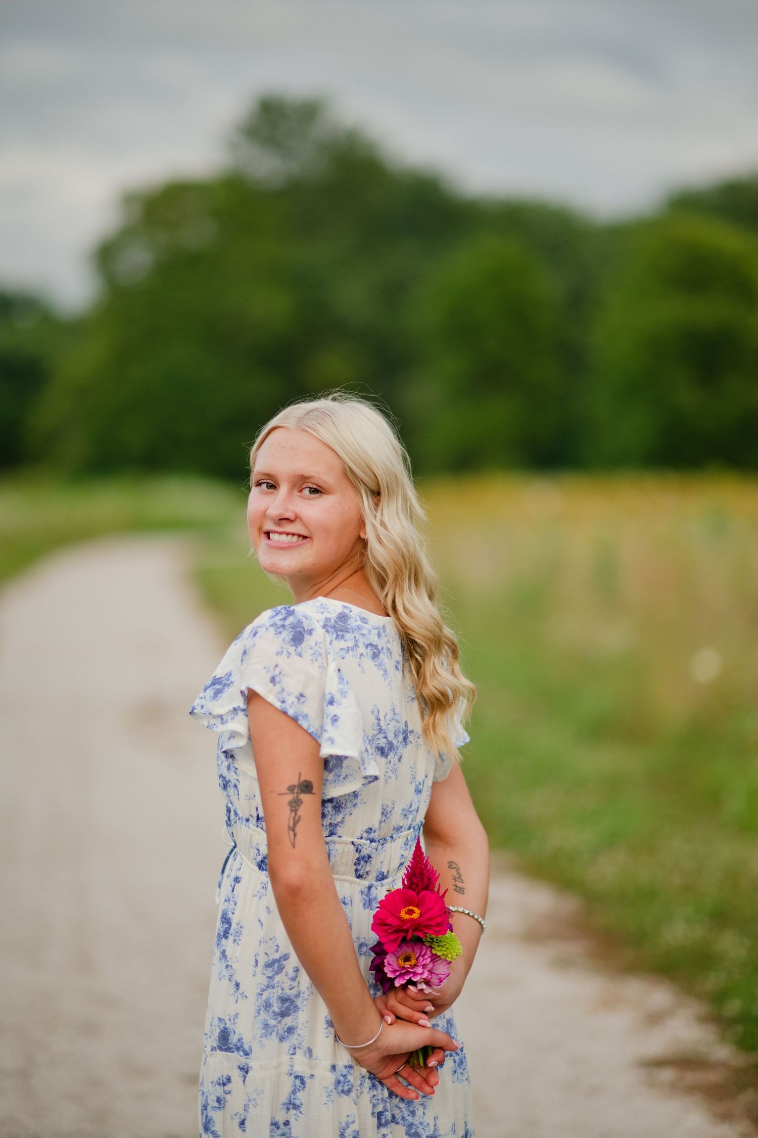 Smiling blonde woman in blue floral dress holds pink flowers on a scenic countryside path.