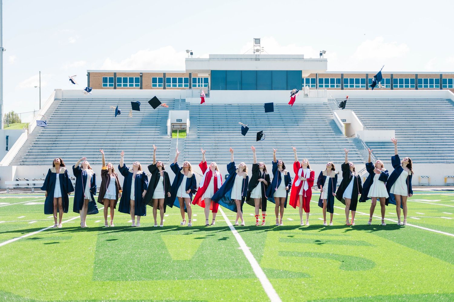 Graduates in navy caps and gowns toss caps in the air on a football field stadium.