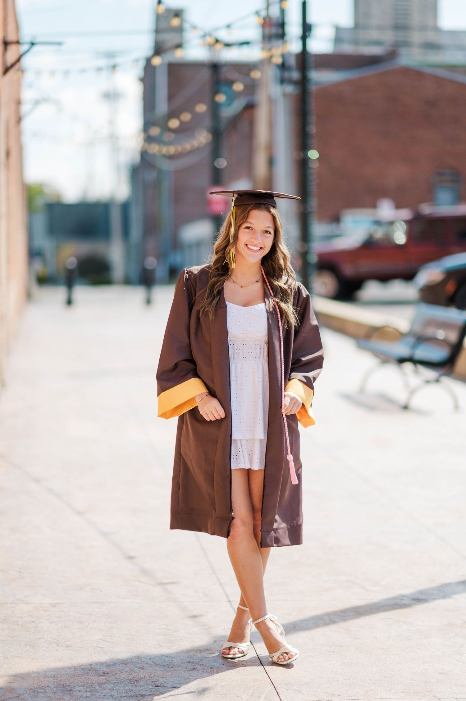 Smiling graduate in brown cap and gown with gold trim walks on sunny urban sidewalk.