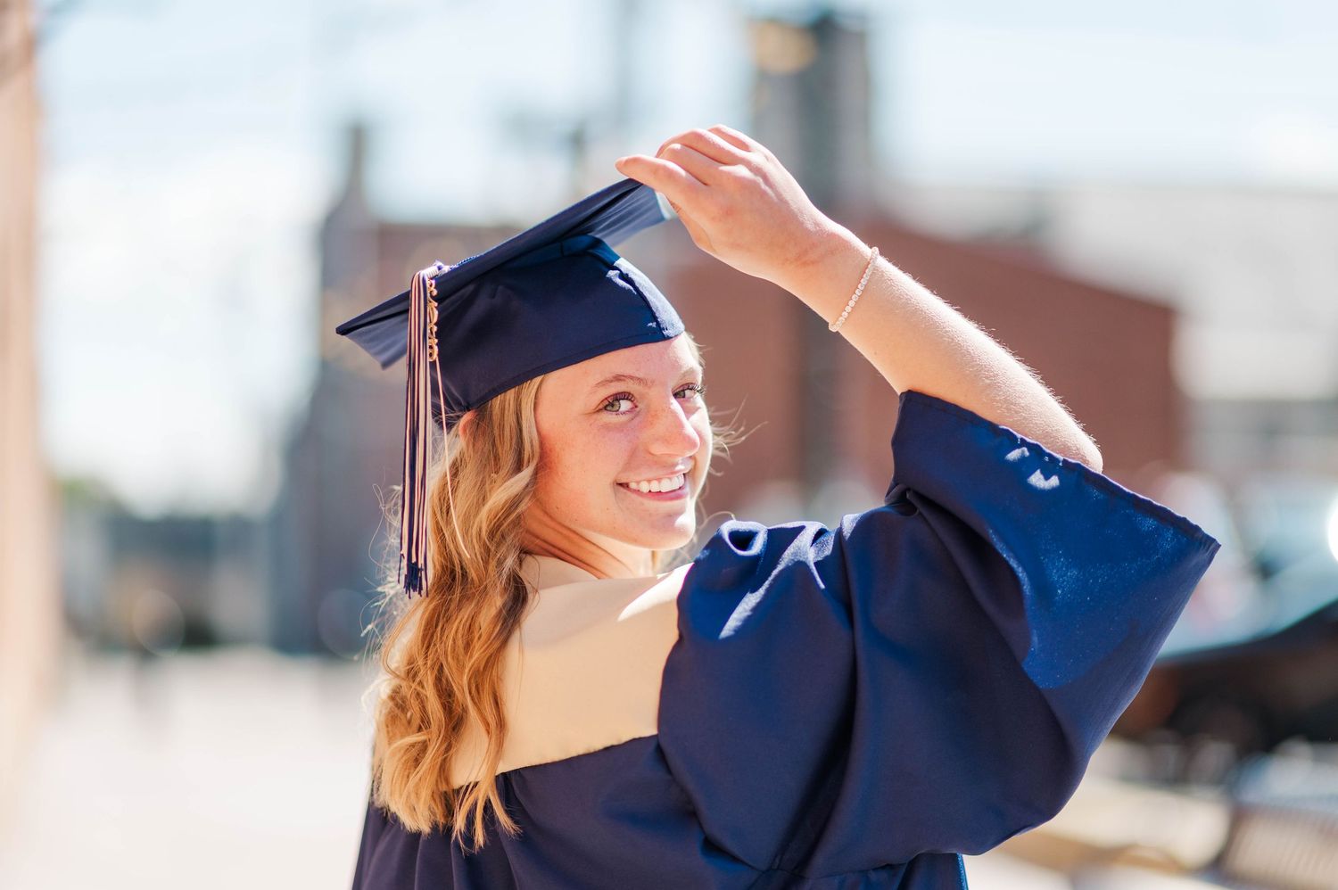 Smiling young woman in navy graduation cap and gown holding her mortarboard outdoors on a sunny day.