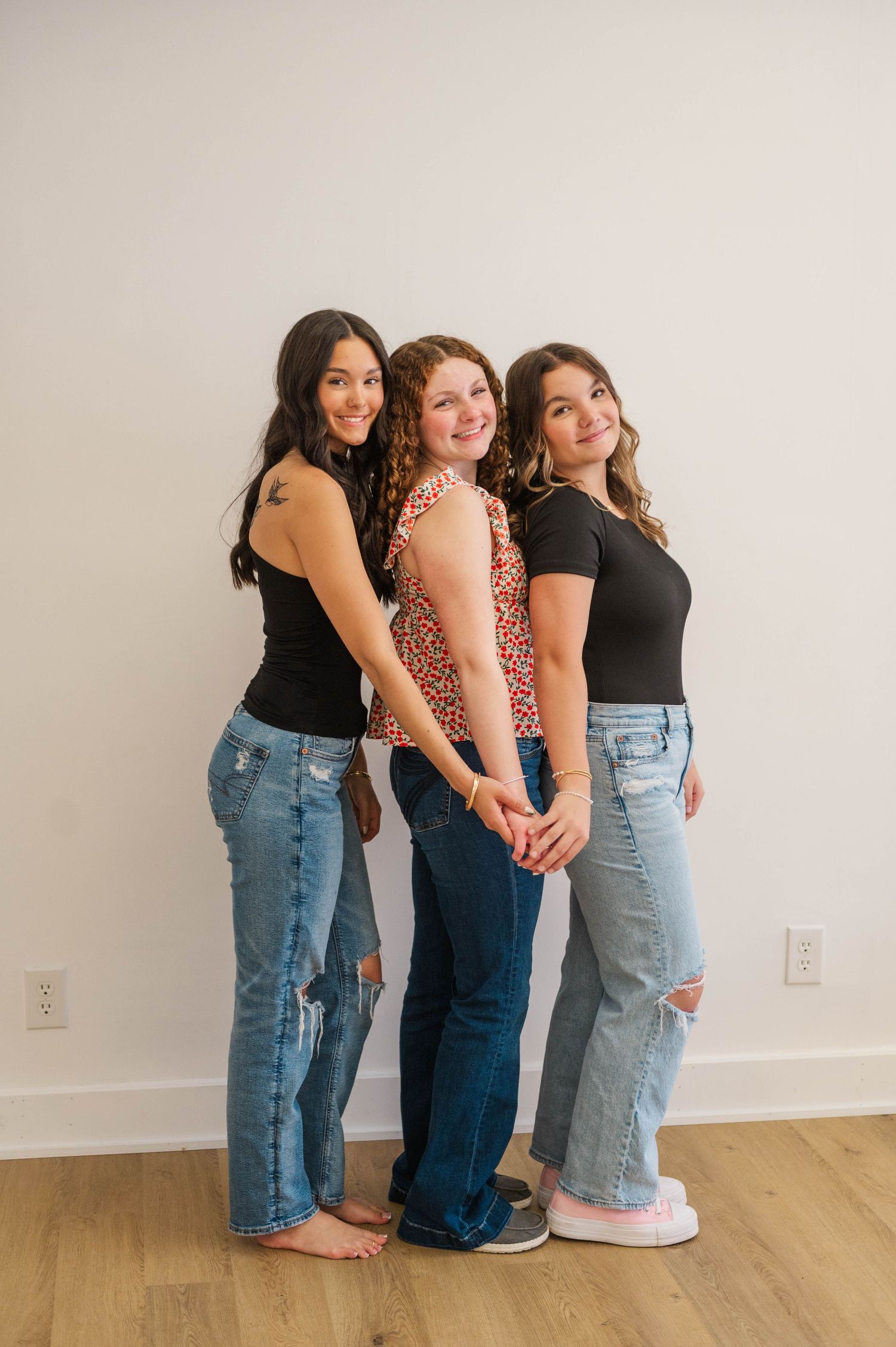 Three young women in jeans pose together against white wall, two in black tops, one in floral.