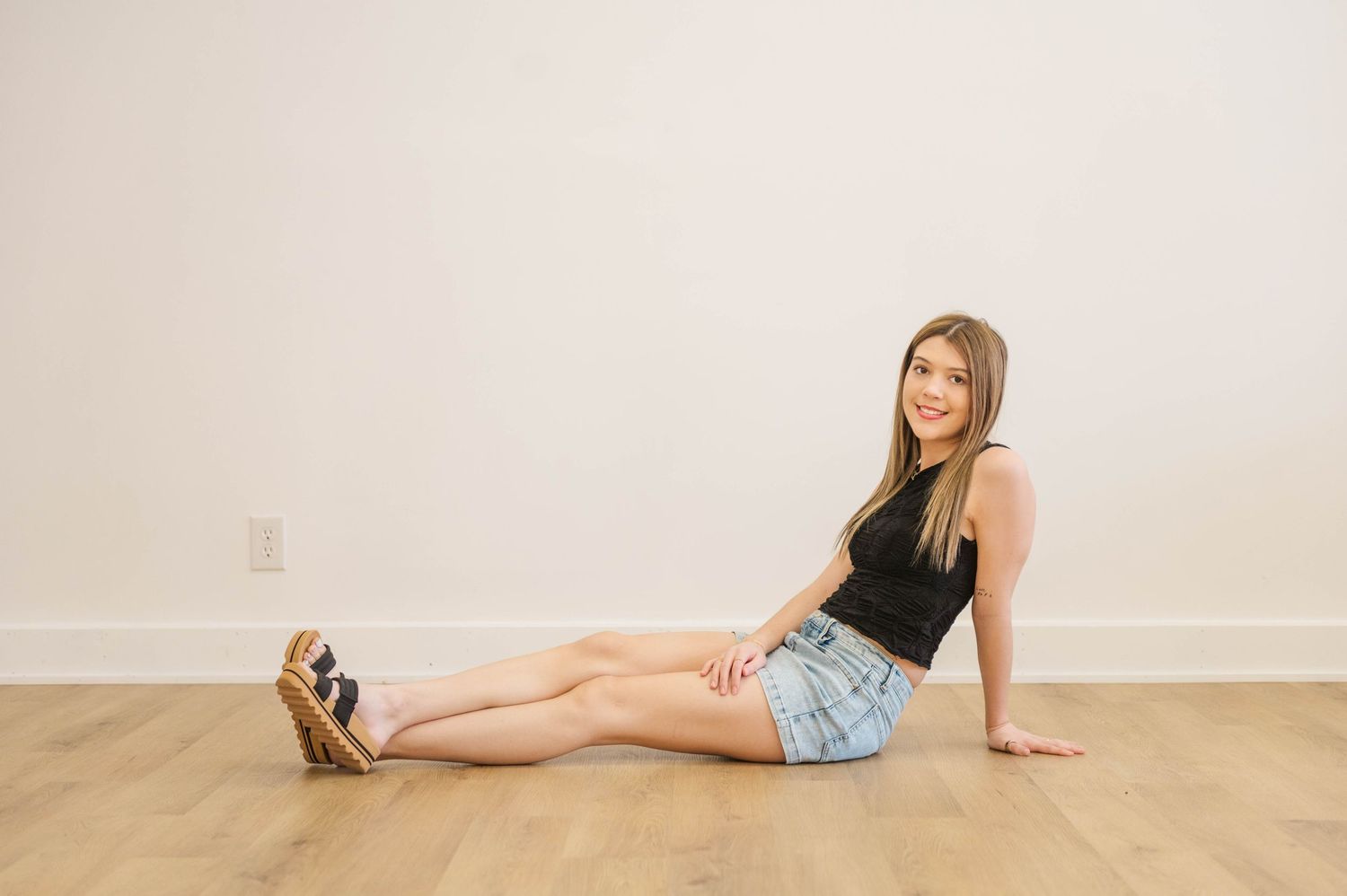 Young woman in black tank and denim shorts sitting on hardwood floor smiling.