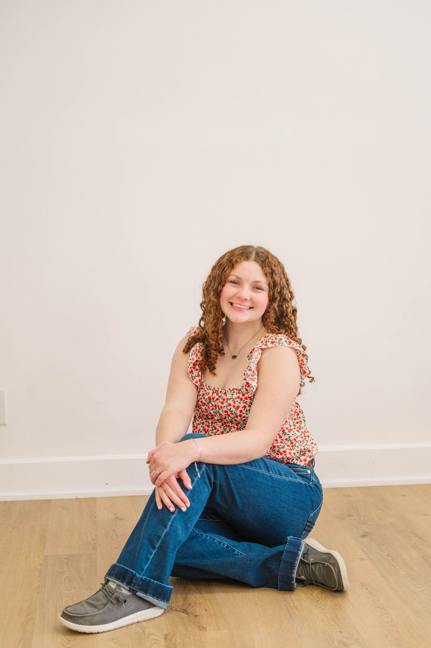 Curly-haired girl in floral tank top and jeans sits cross-legged on hardwood floor, smiling brightly.