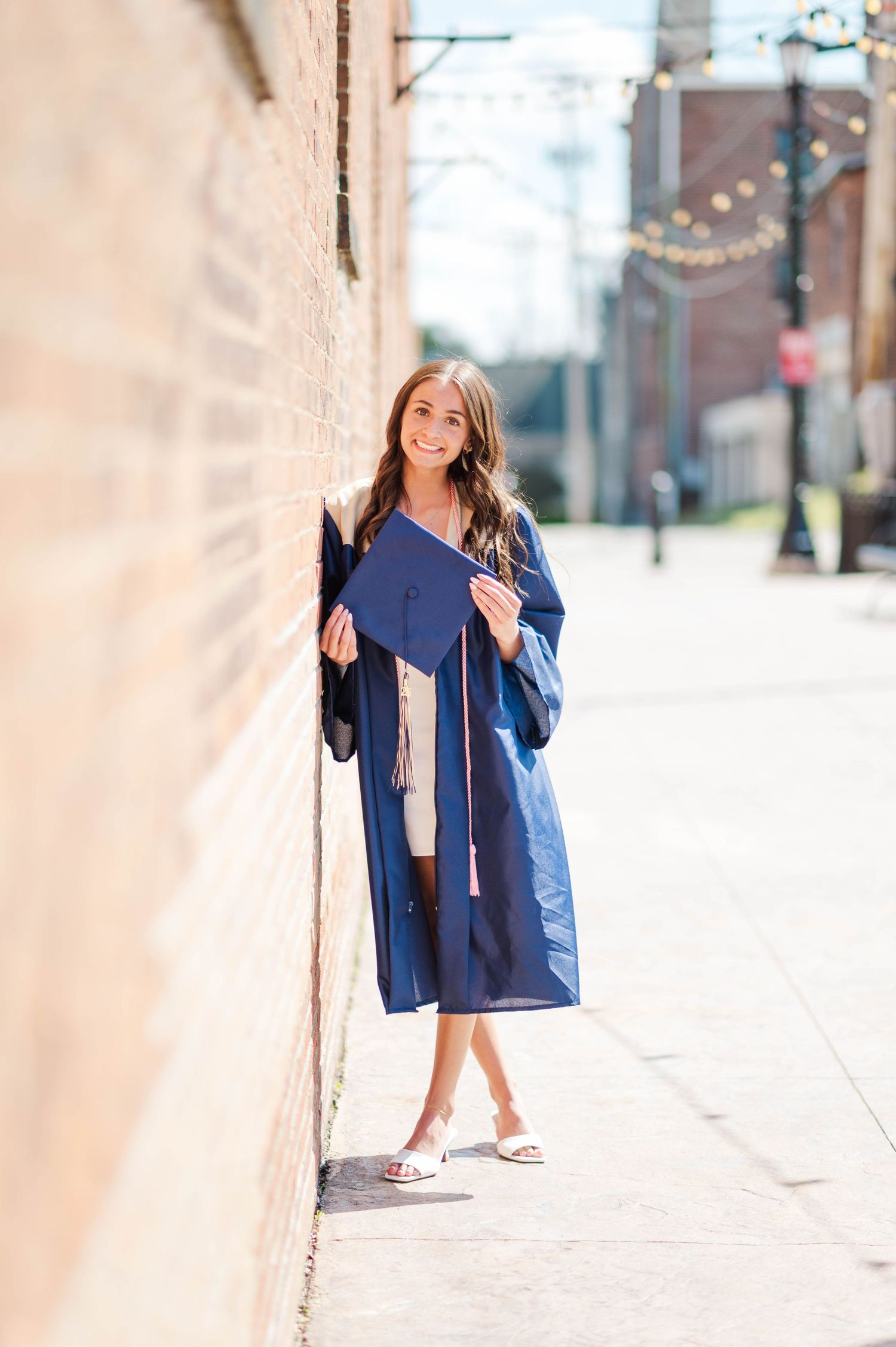 Smiling brunette in navy graduation gown leaning against brick wall on sunny sidewalk.
