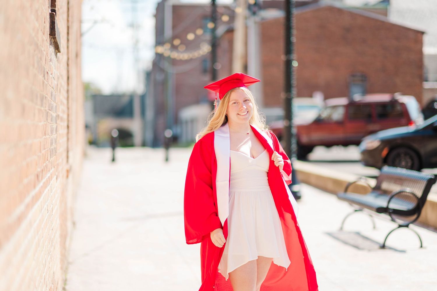 Happy graduate in red cap and gown walking along a brick-lined urban street.