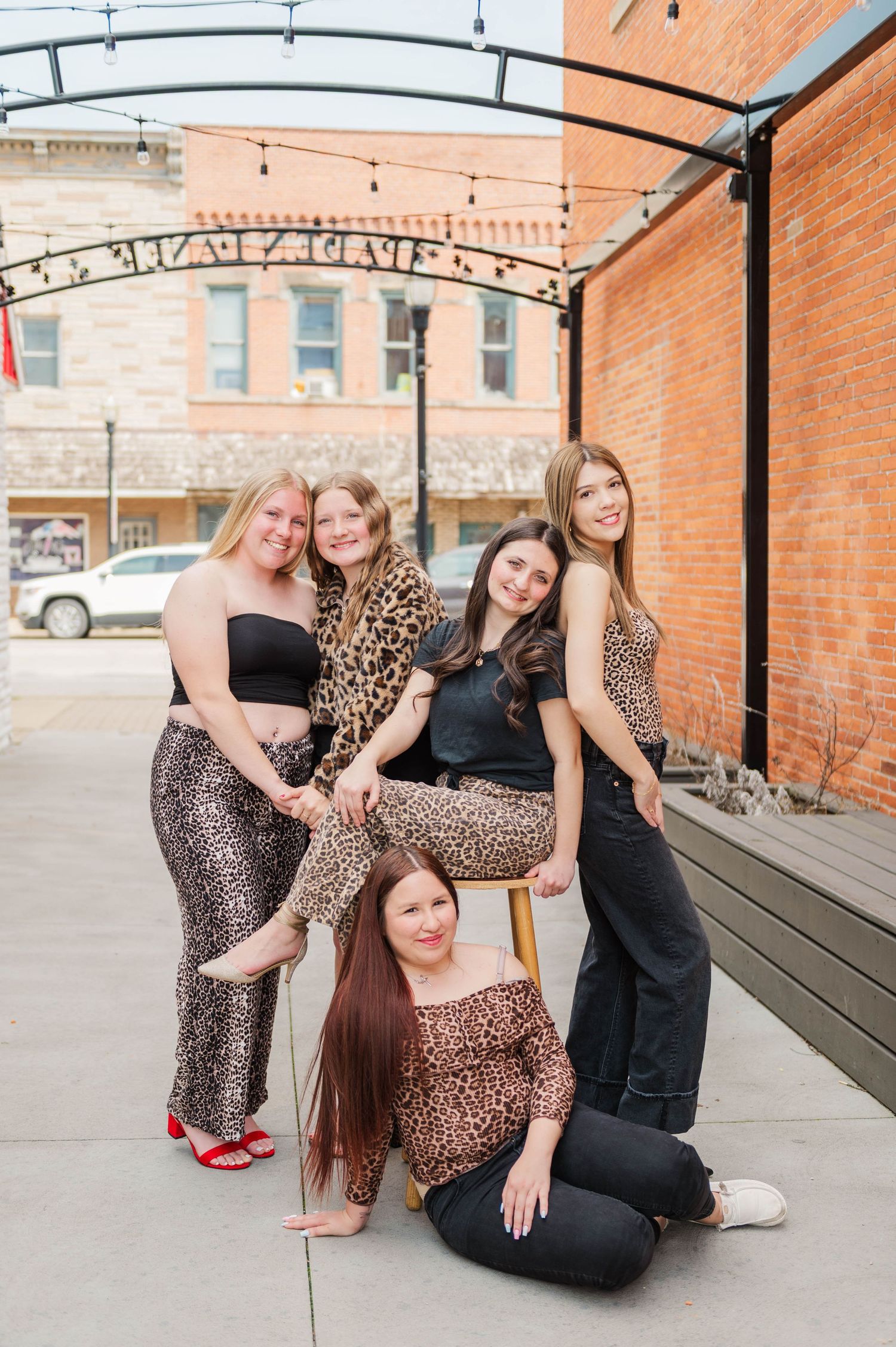 Group of five young women pose together near stool under string lights against brick building backdrop.