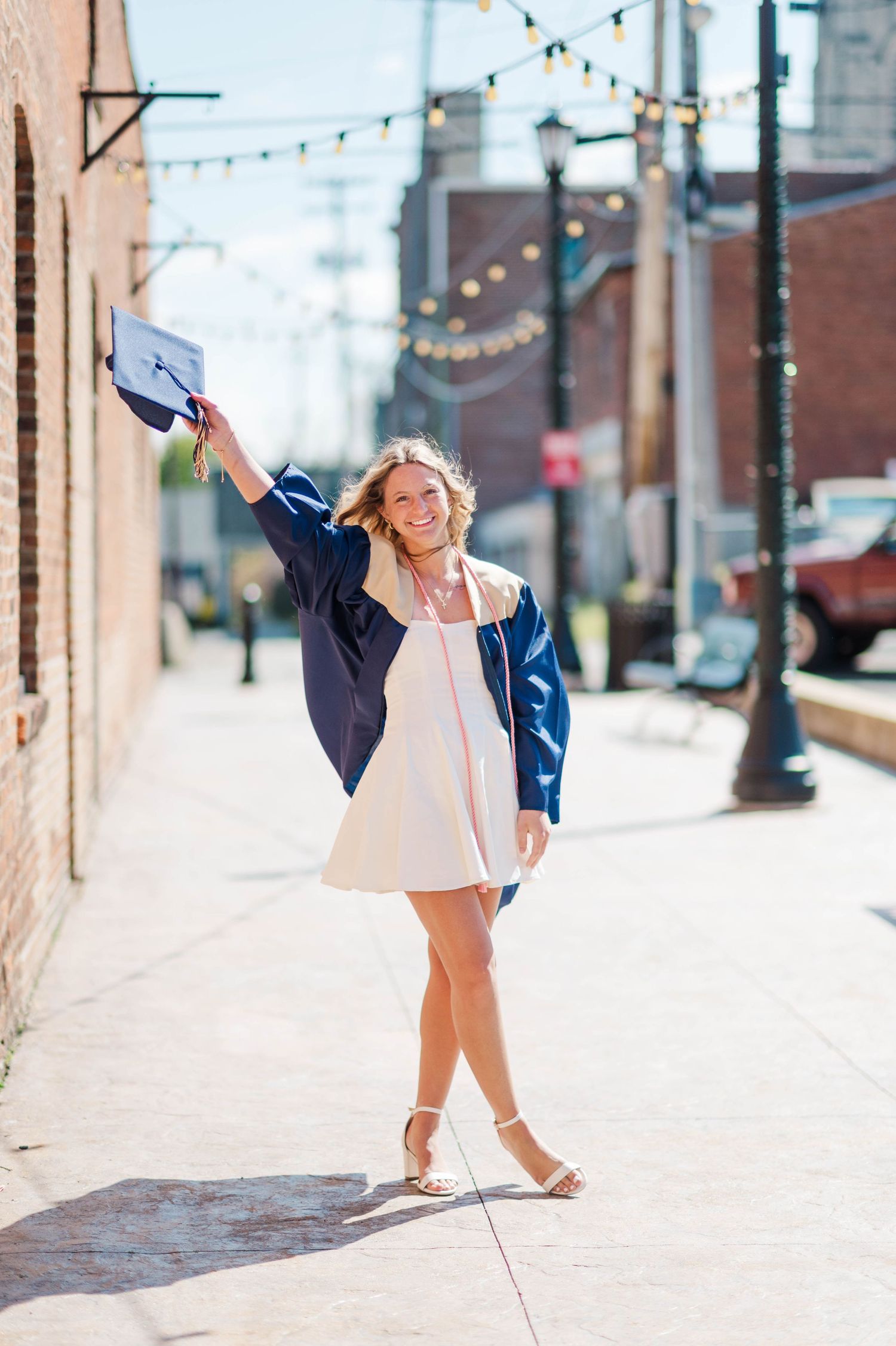 Smiling graduate in navy cap and gown over white dress celebrates on sunny urban brick alley sidewalk.