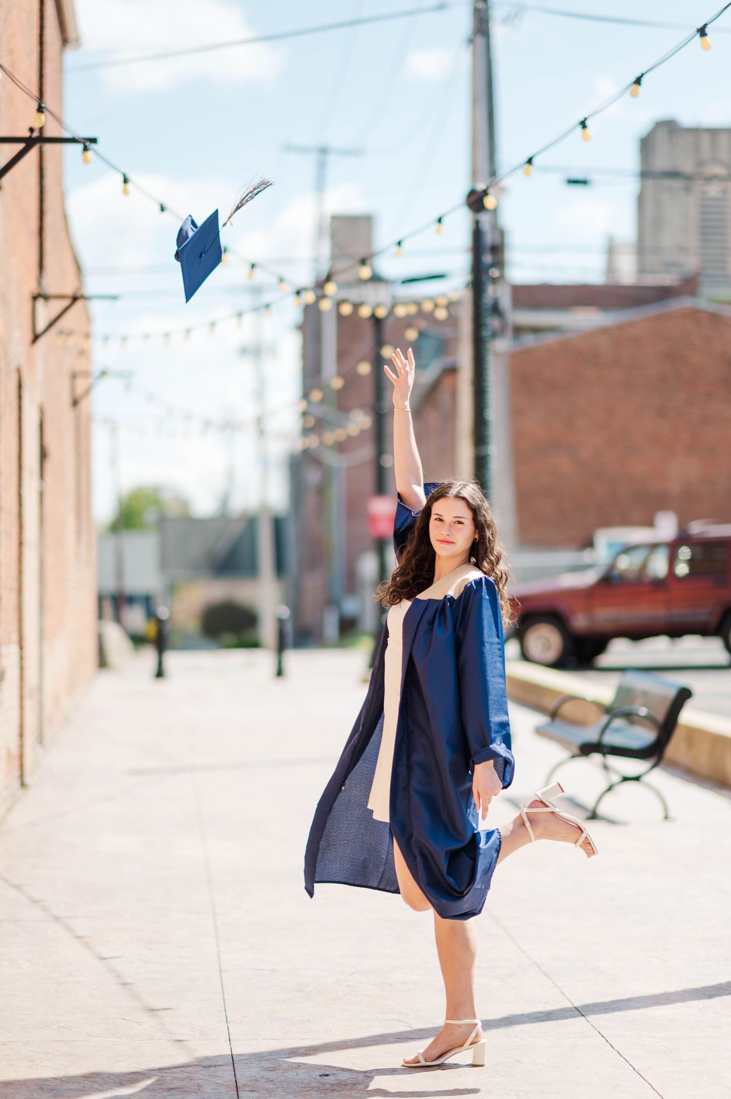 Joyful graduate in navy gown tosses her cap skyward on a sunny urban sidewalk.