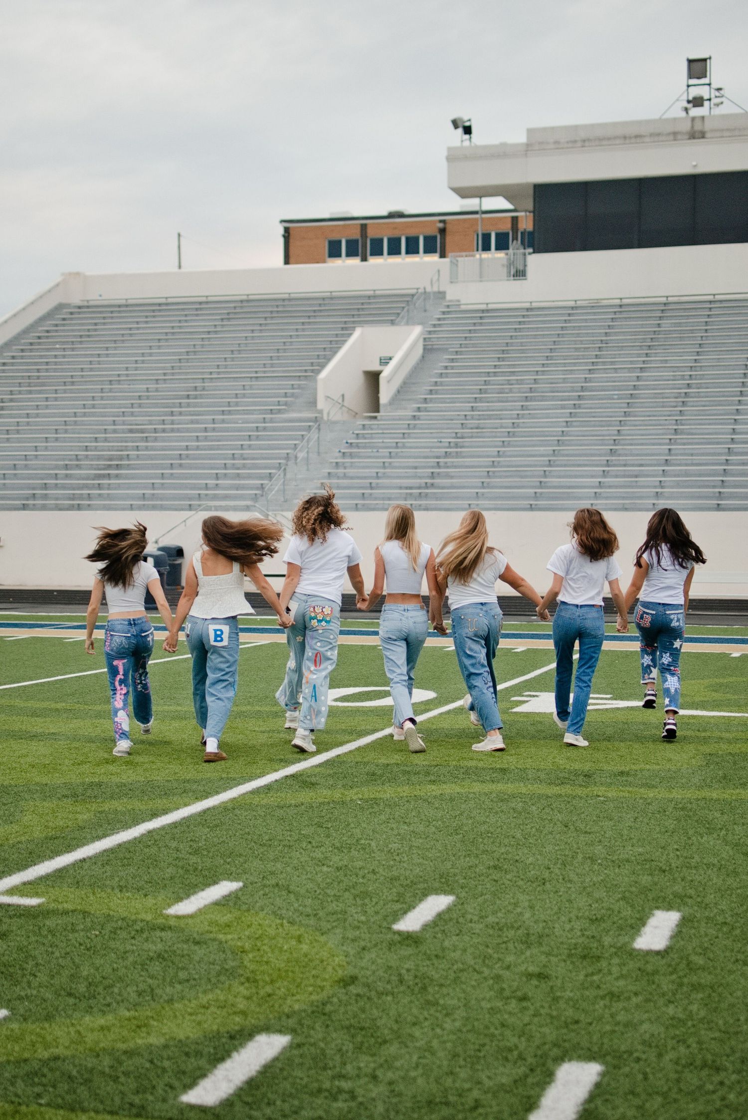 Group of seven girls in white tops and painted jeans run holding hands across a football field.