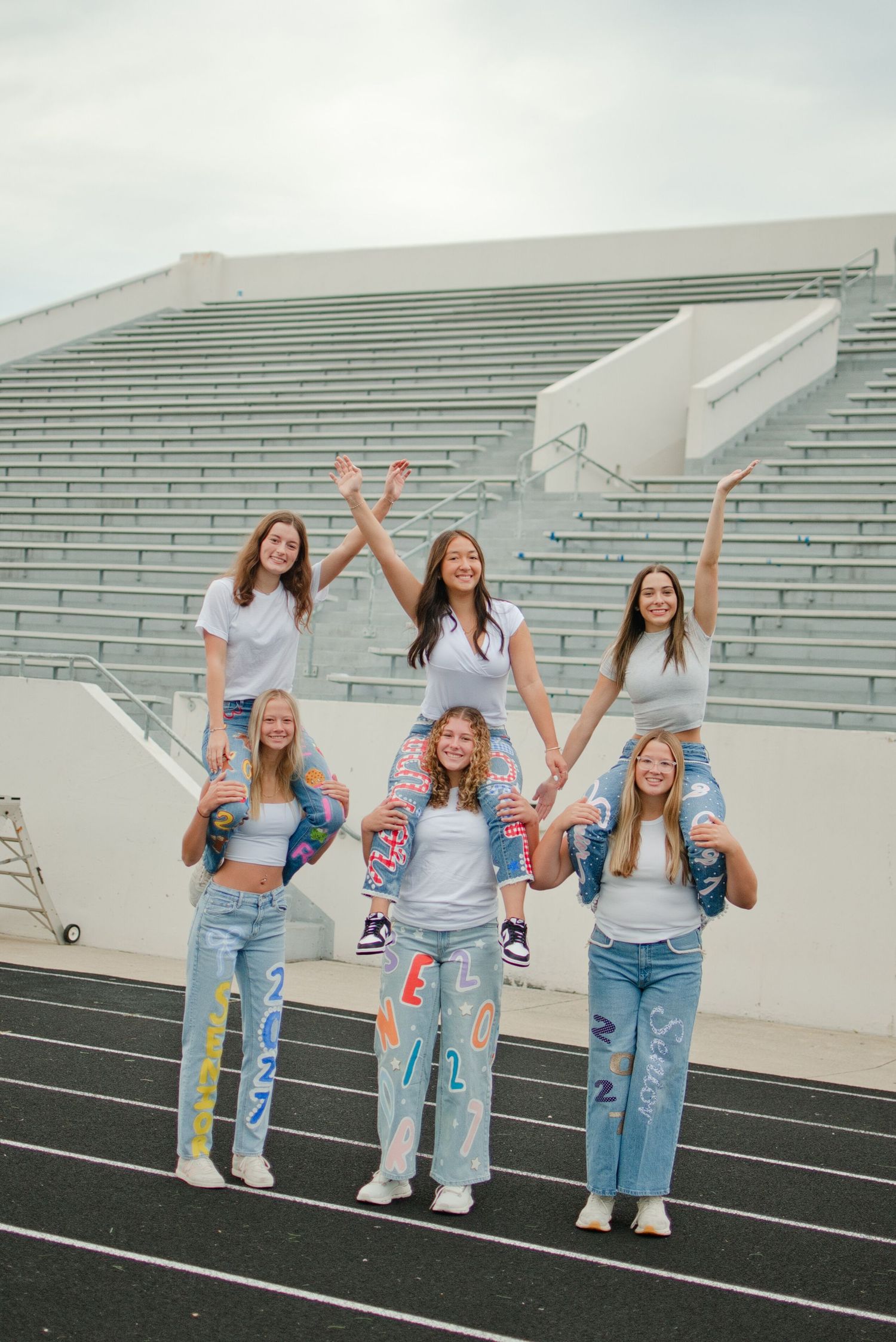 Six teenage girls in white tees and painted jeans pose in a cheerful pyramid on a stadium track.