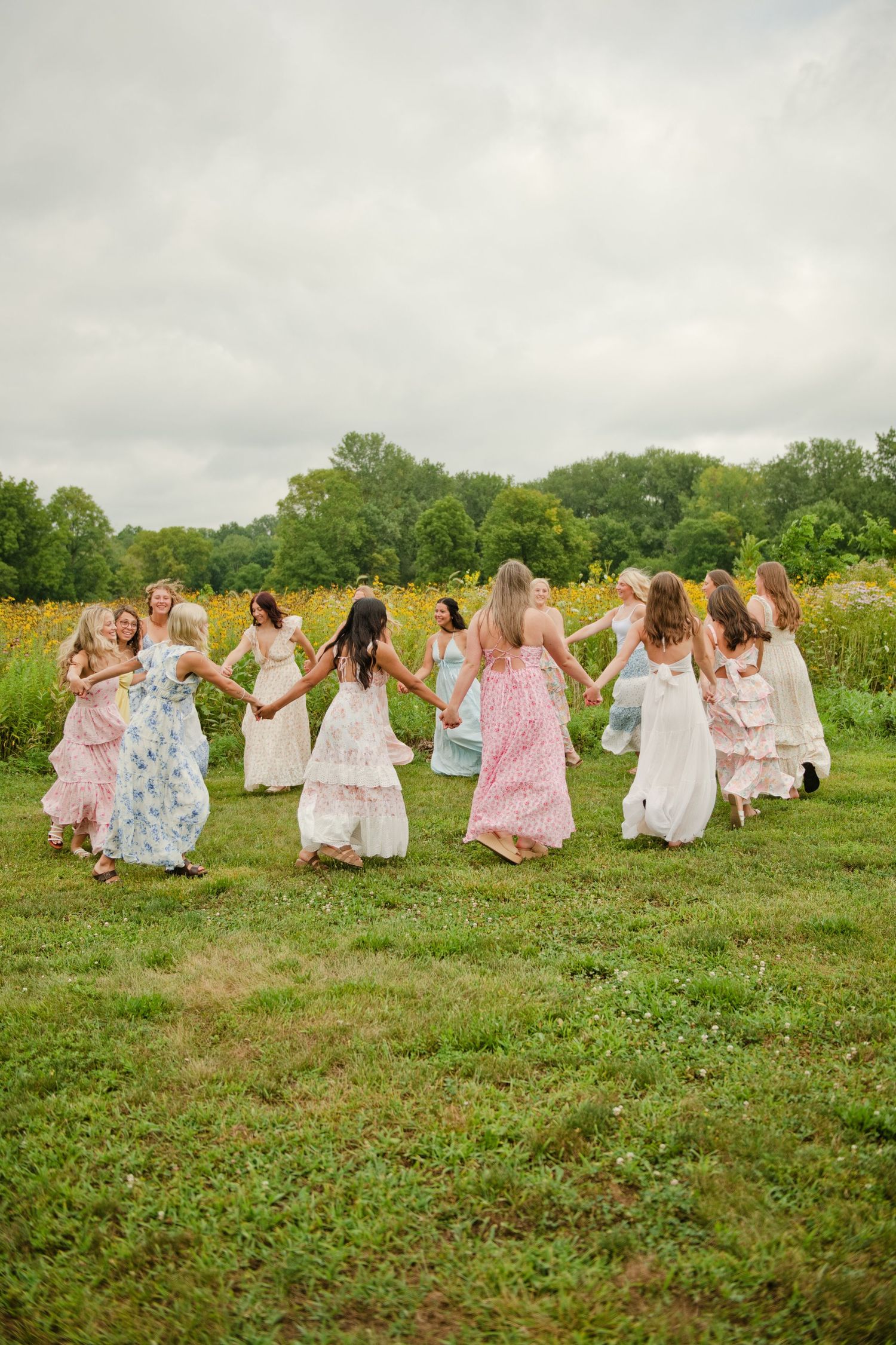 Bridesmaids in colorful floral maxi dresses hold hands in a circle dancing in a green field.