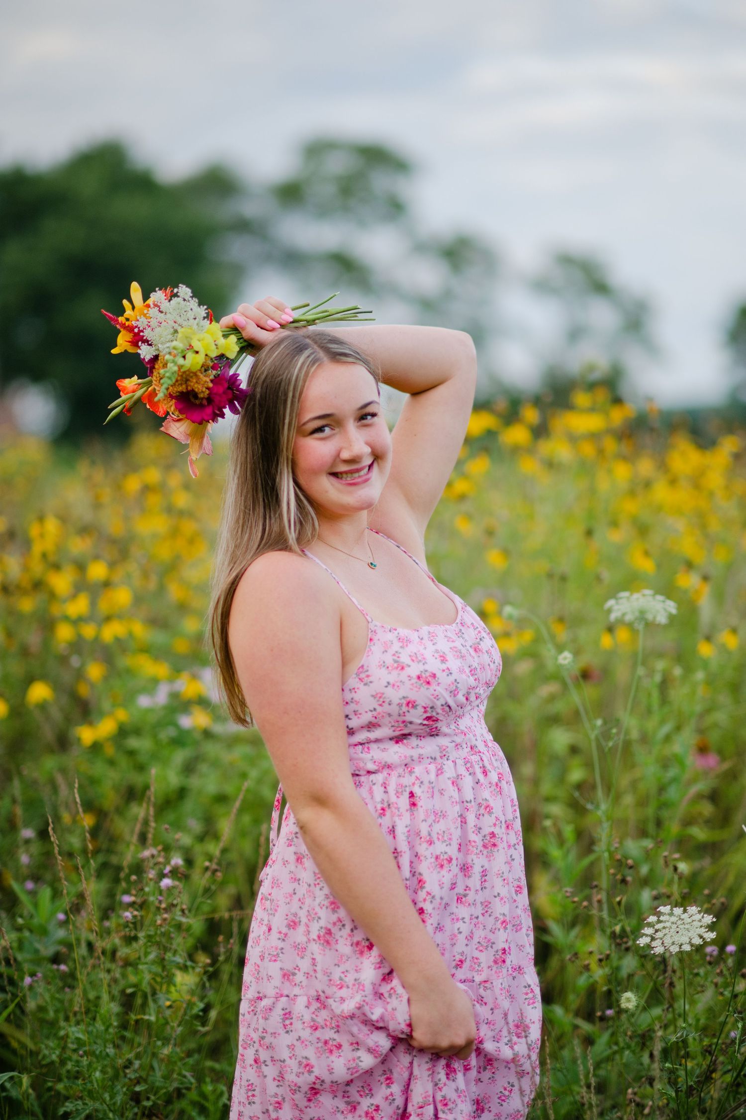 Smiling young woman in pink floral dress holds wildflower bouquet in a blooming meadow.