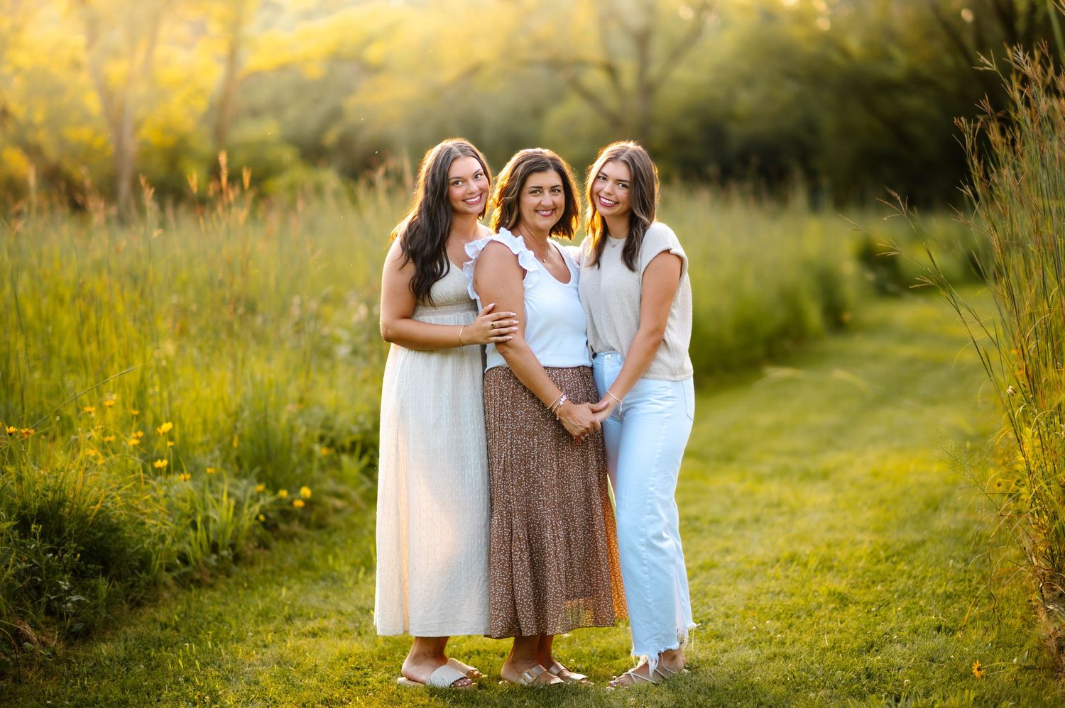 Three people share laughter and hugs while standing on a grassy path at sunset.