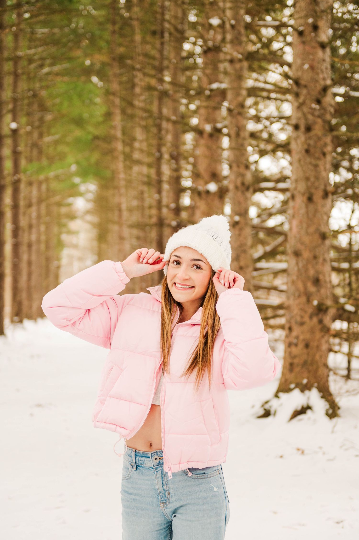 Winter fashion shoot featuring pink puffer jacket and light denim in snowy forest setting.