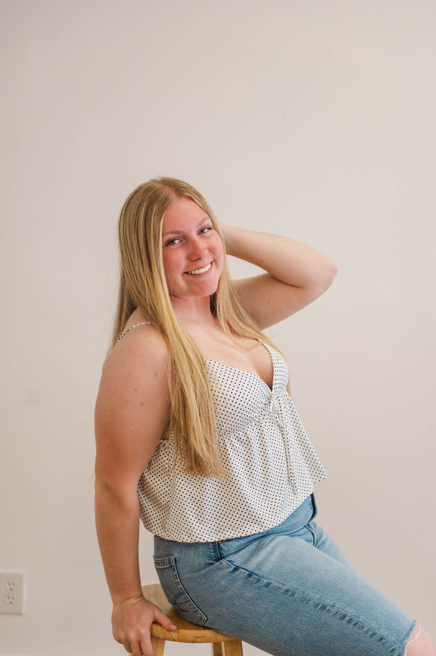 Smiling blonde woman sits on wooden stool with hand in hair, wearing polka dot top and jeans.