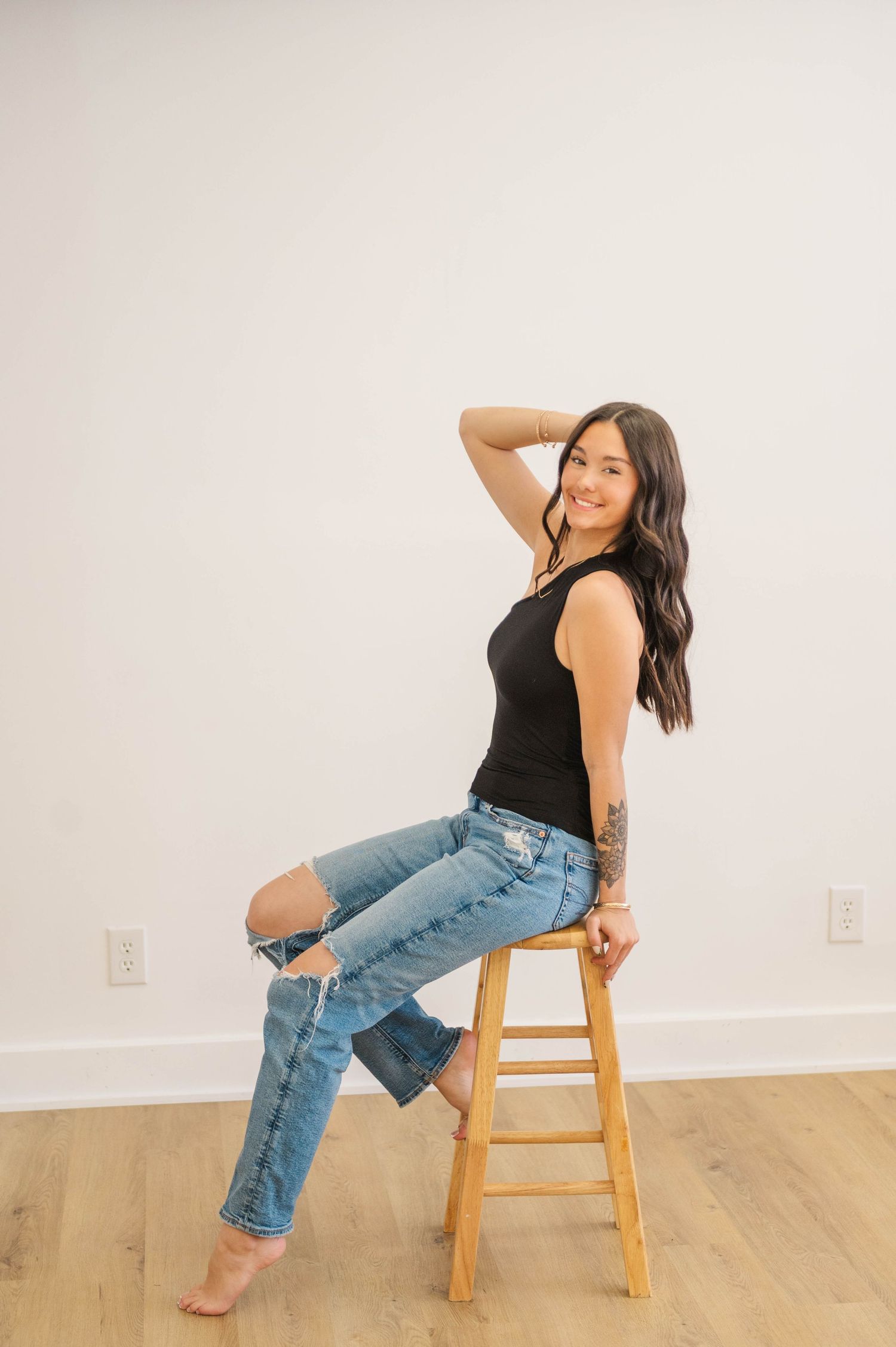 Woman with tattoos sits on wooden stool in black tank top and ripped jeans, smiling over shoulder.